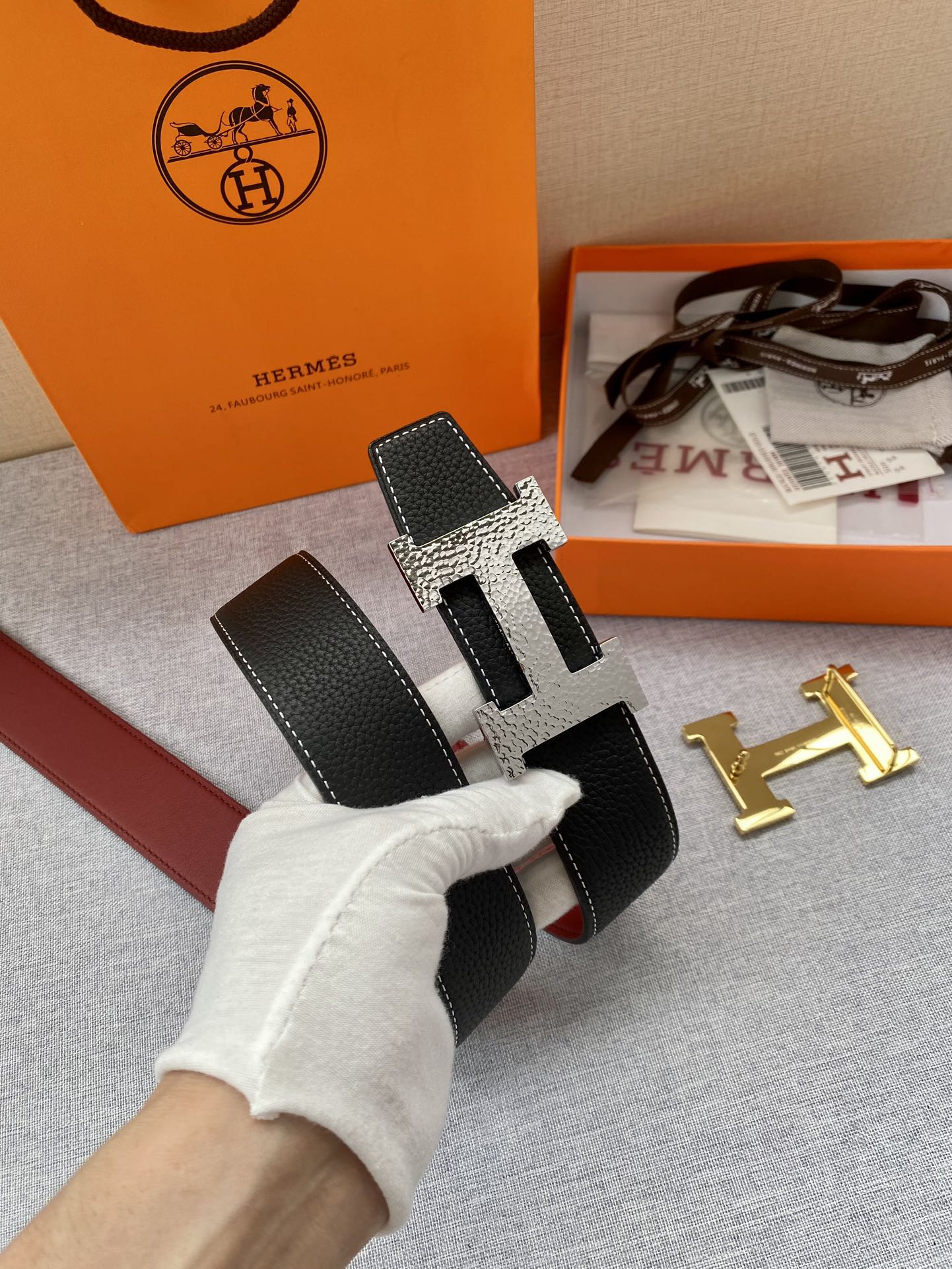 Hermes belt with black textured leather and gold-tone H buckle, shown at a diagonal angle.