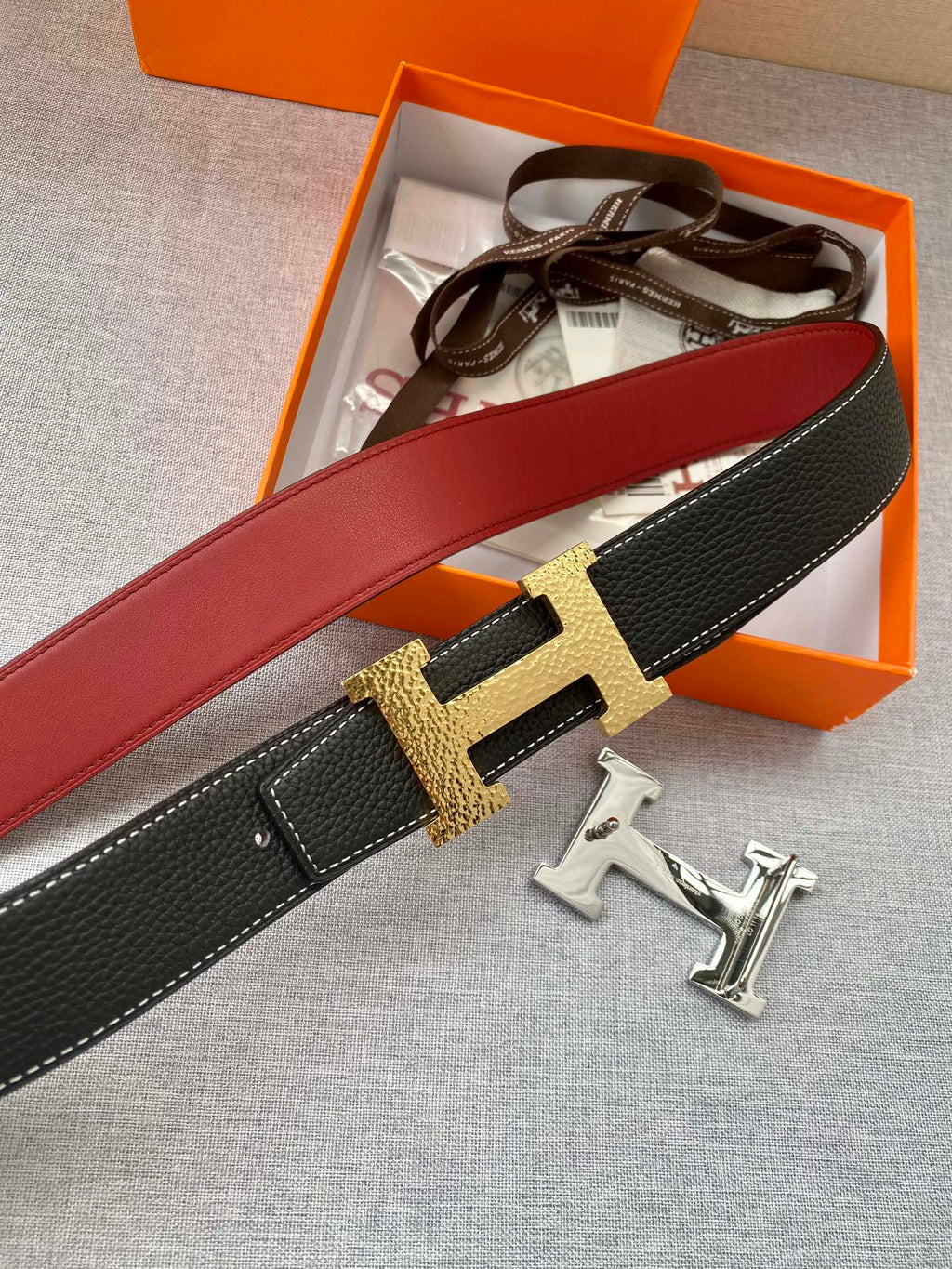 Hermes belt close-up with gold-tone H buckle, black textured leather and red strap side.