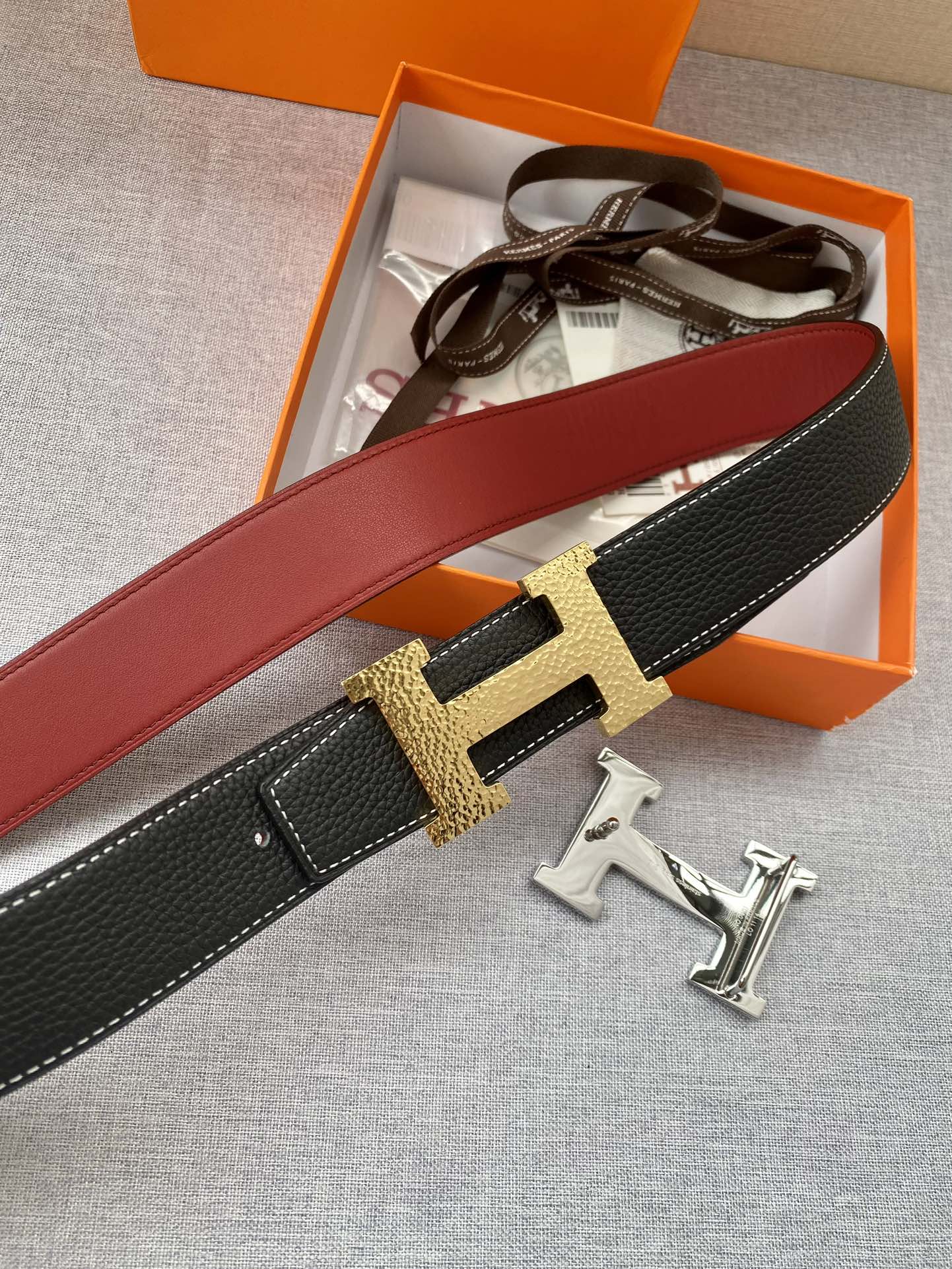 Hermes belt close-up with gold-tone H buckle, black textured leather and red strap side.