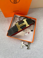 Hermes belt in box: black textured strap with gold-tone H buckle and red underside detail.