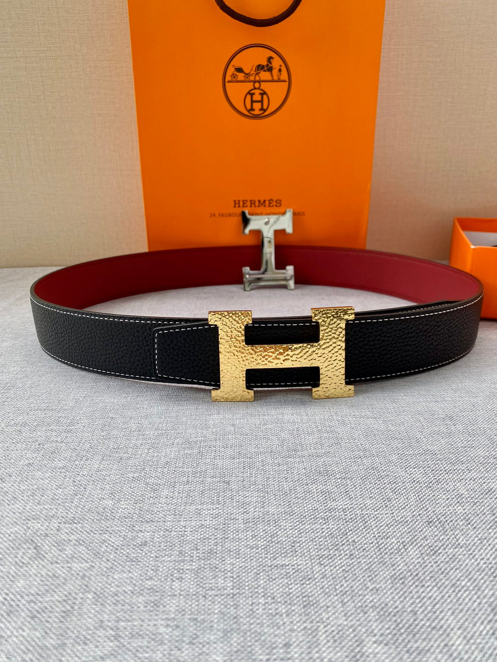 Hermes belt on flat surface: black textured strap with red interior and gold-tone H buckle.