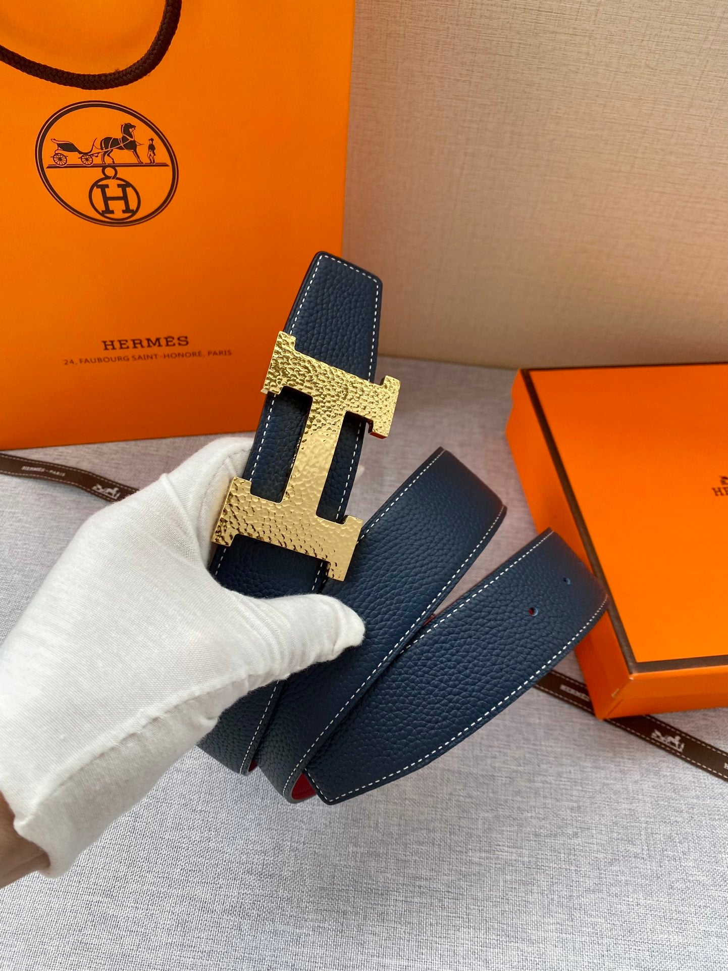 Hermes dark blue leather belt with gold H buckle, held by a gloved hand.