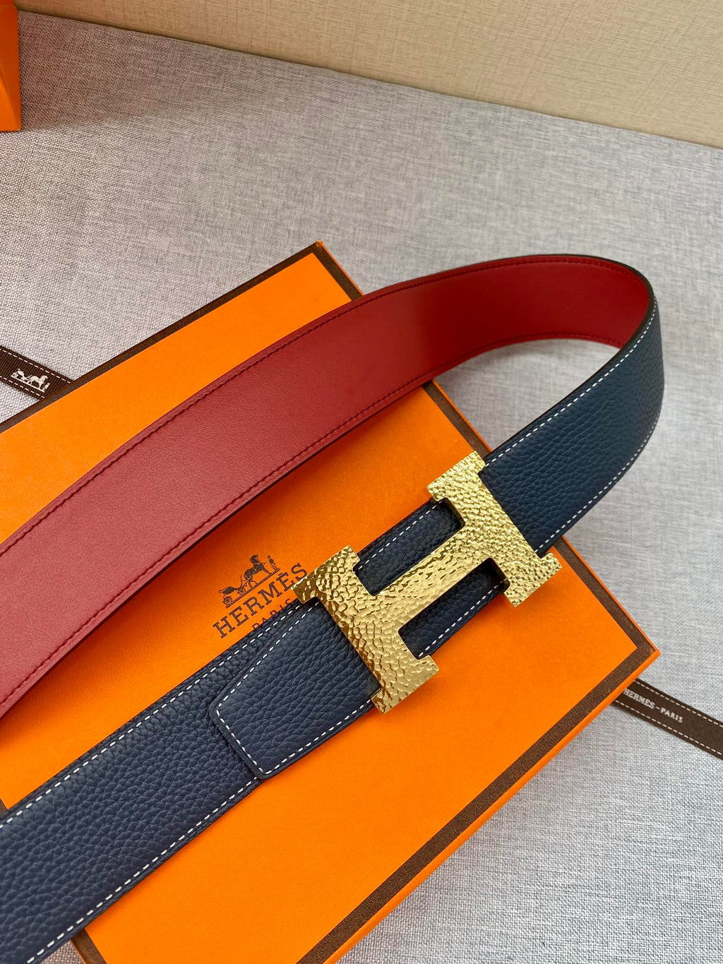 Hermes dark blue leather belt close-up with gold H buckle and red lining edge.