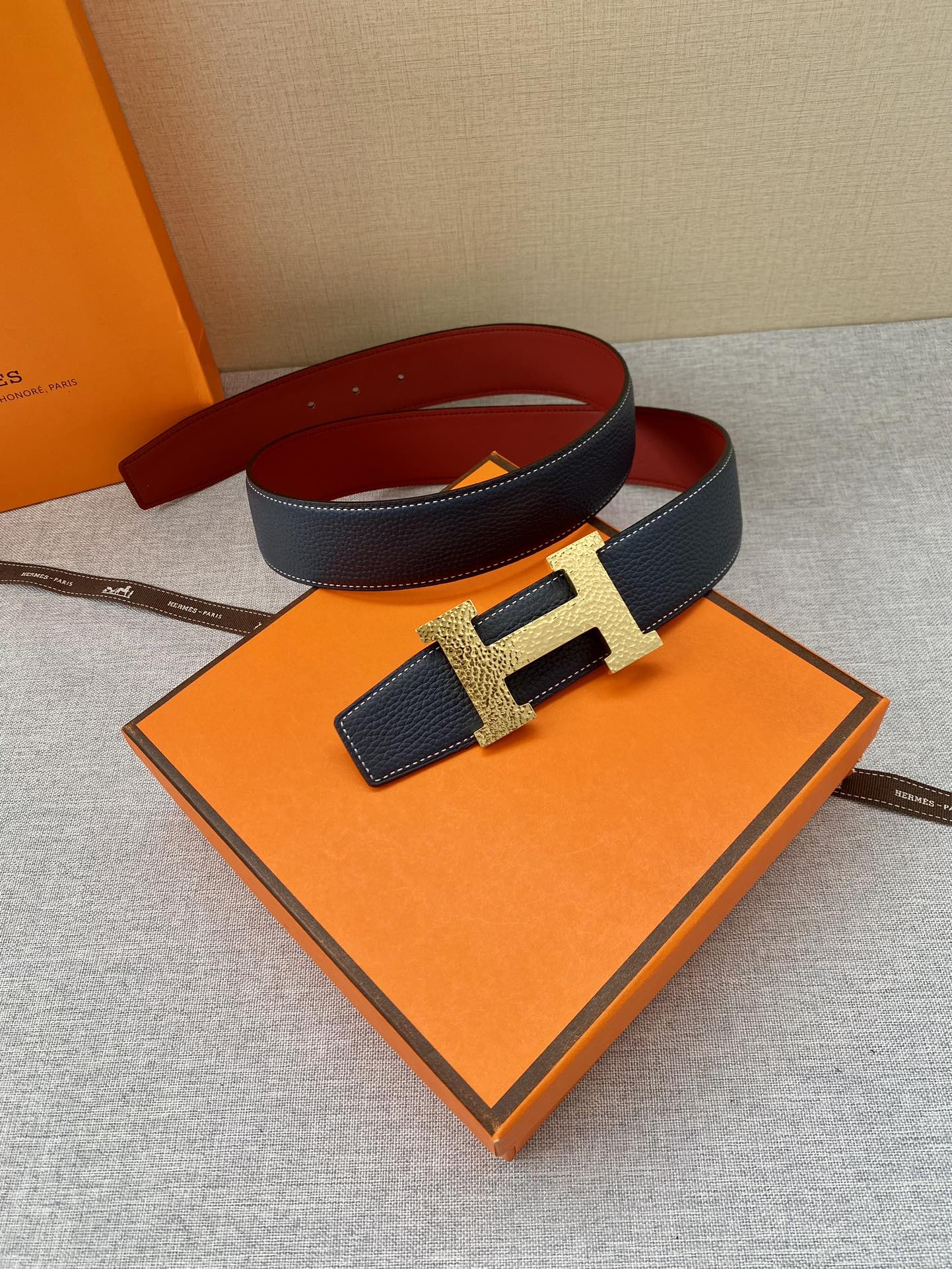 Navy leather belt with gold-tone H buckle, folded beside orange box