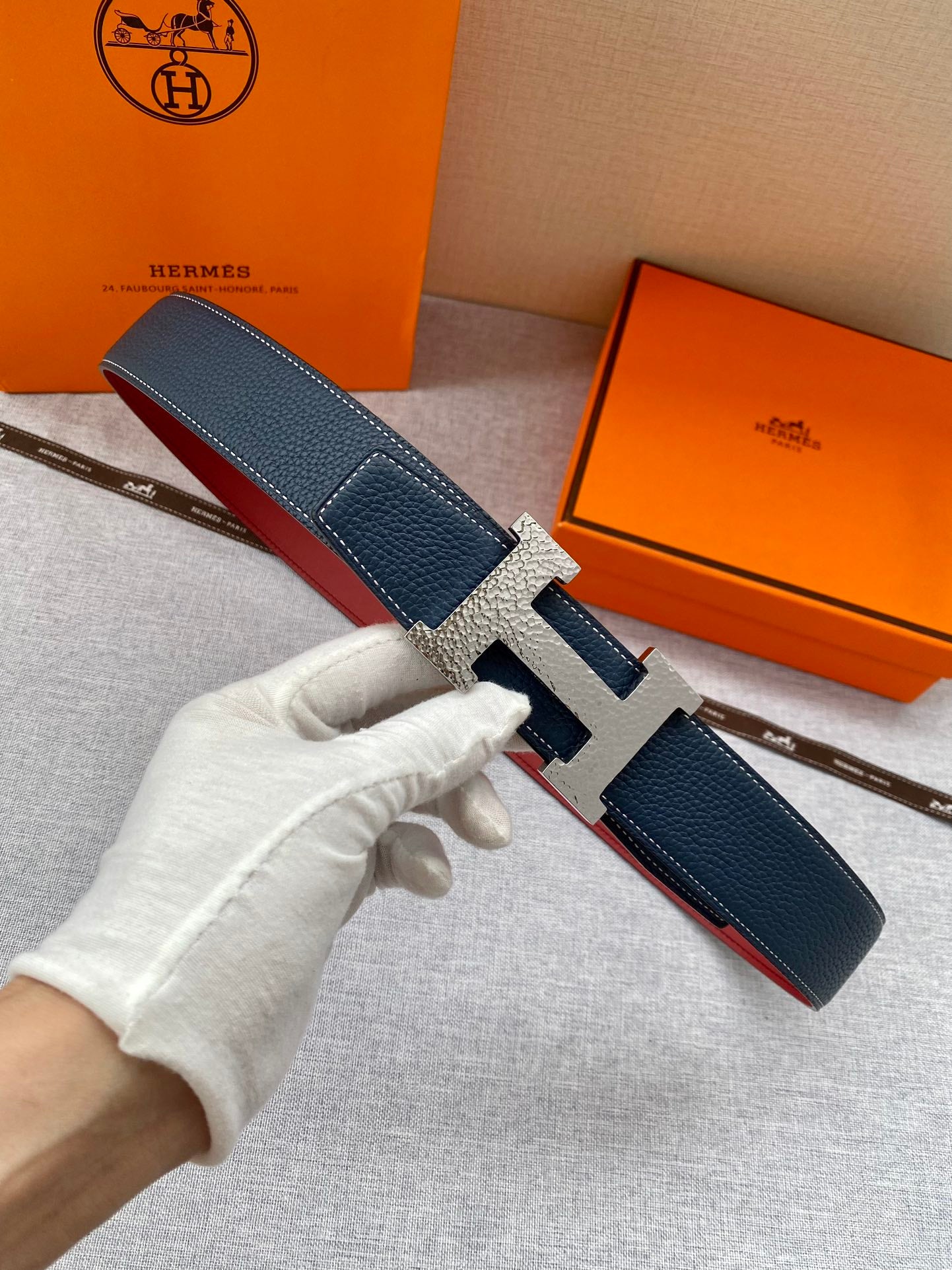 Hermes belt in dark blue leather with silver H buckle, held by a gloved hand.