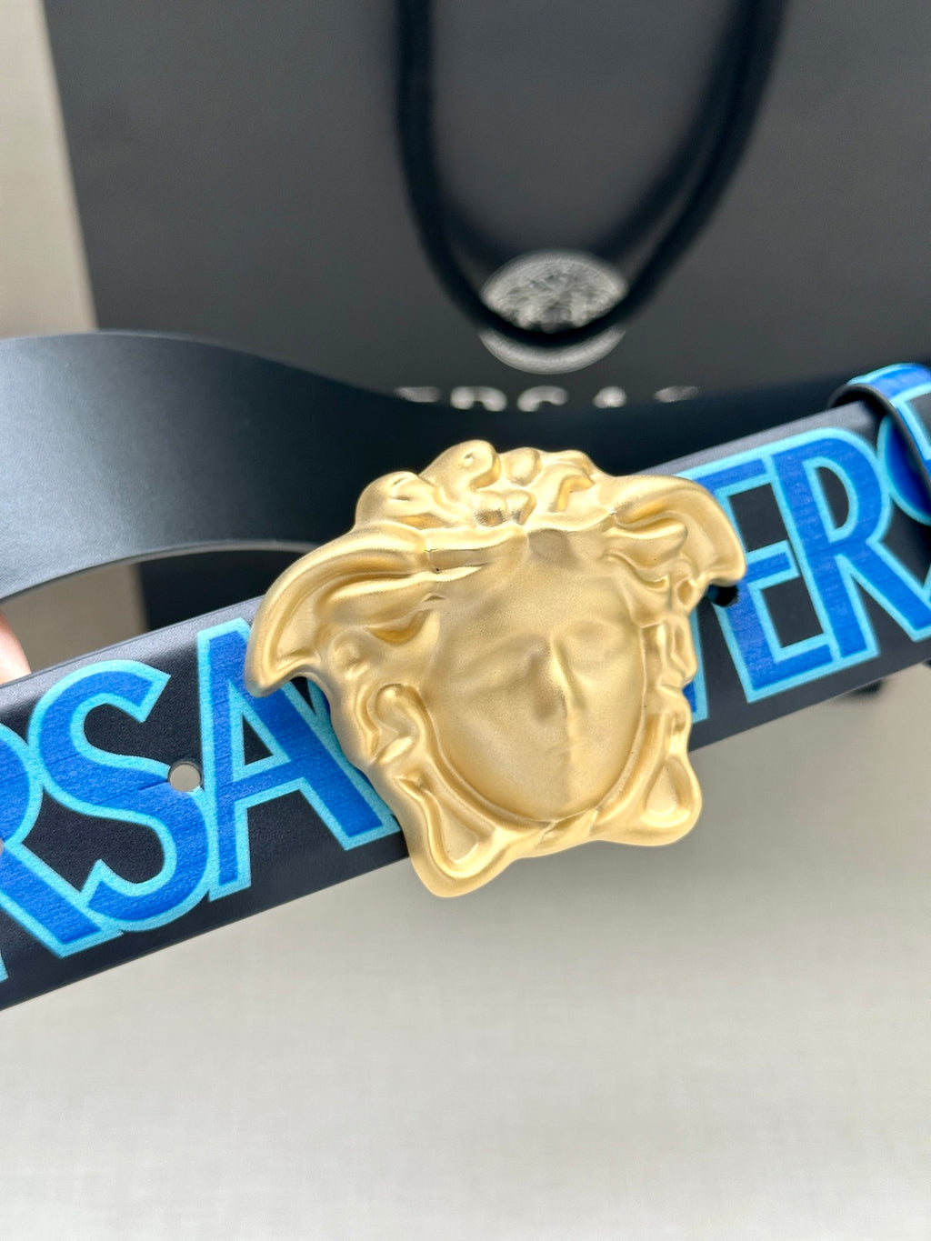 Versace belt buckle close-up: gold Medusa on dark blue strap with blue logo print.