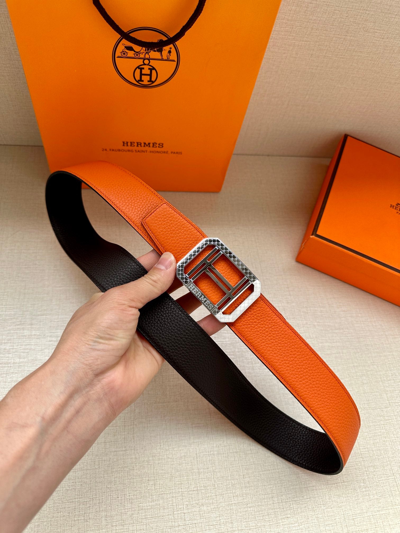 Hermes belt laid flat: orange leather strap with silver square buckle and black reverse side.