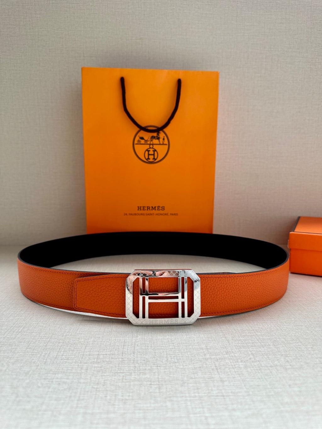 Hermes belt with orange leather strap and centered silver buckle, black underside visible.