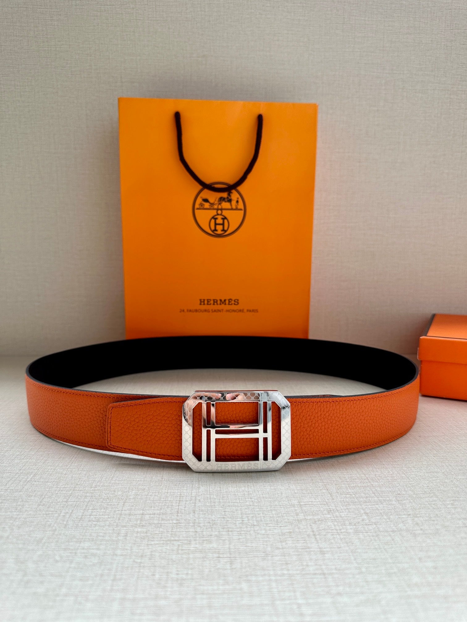 Hermes belt with orange leather strap and centered silver buckle, black underside visible.