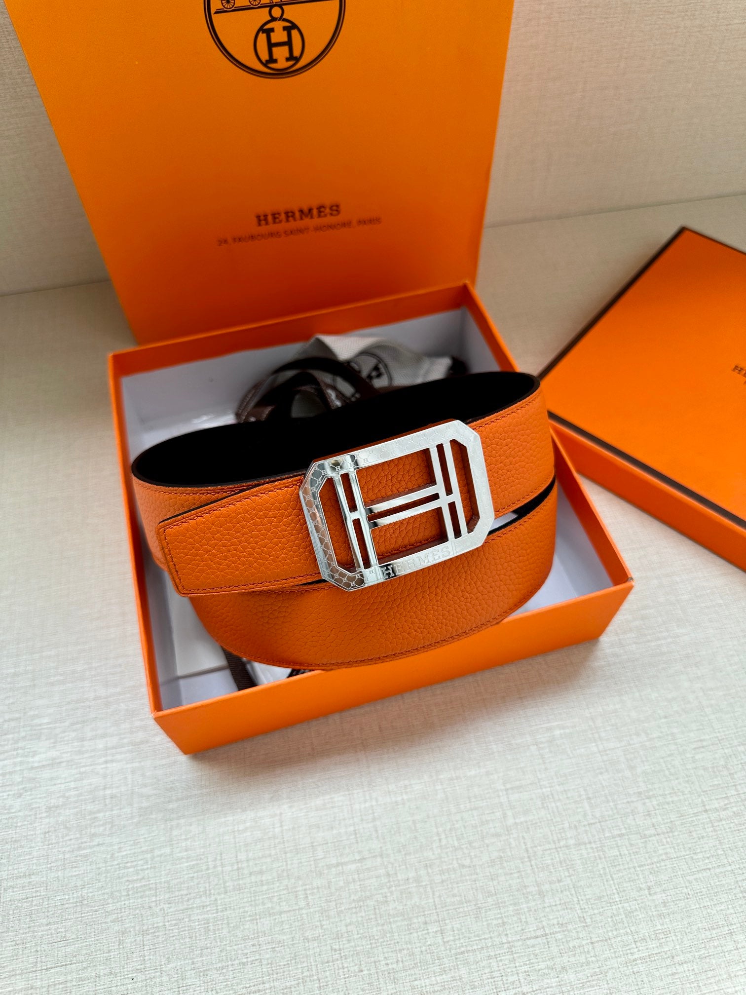 Hermes belt in packaging: orange leather strap with silver buckle and black underside.