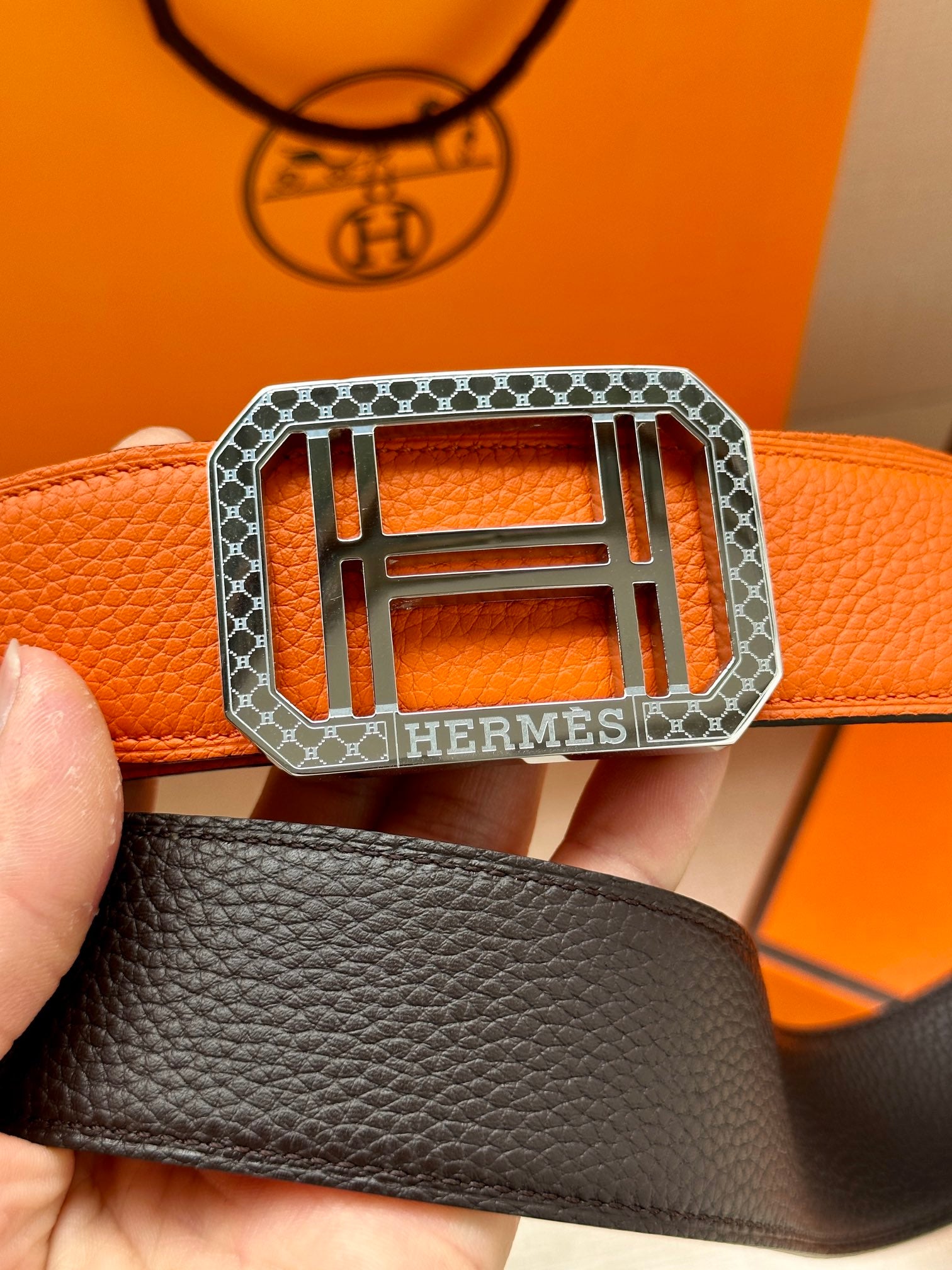 Hermes belt buckle close-up: silver H hardware on orange textured leather strap.