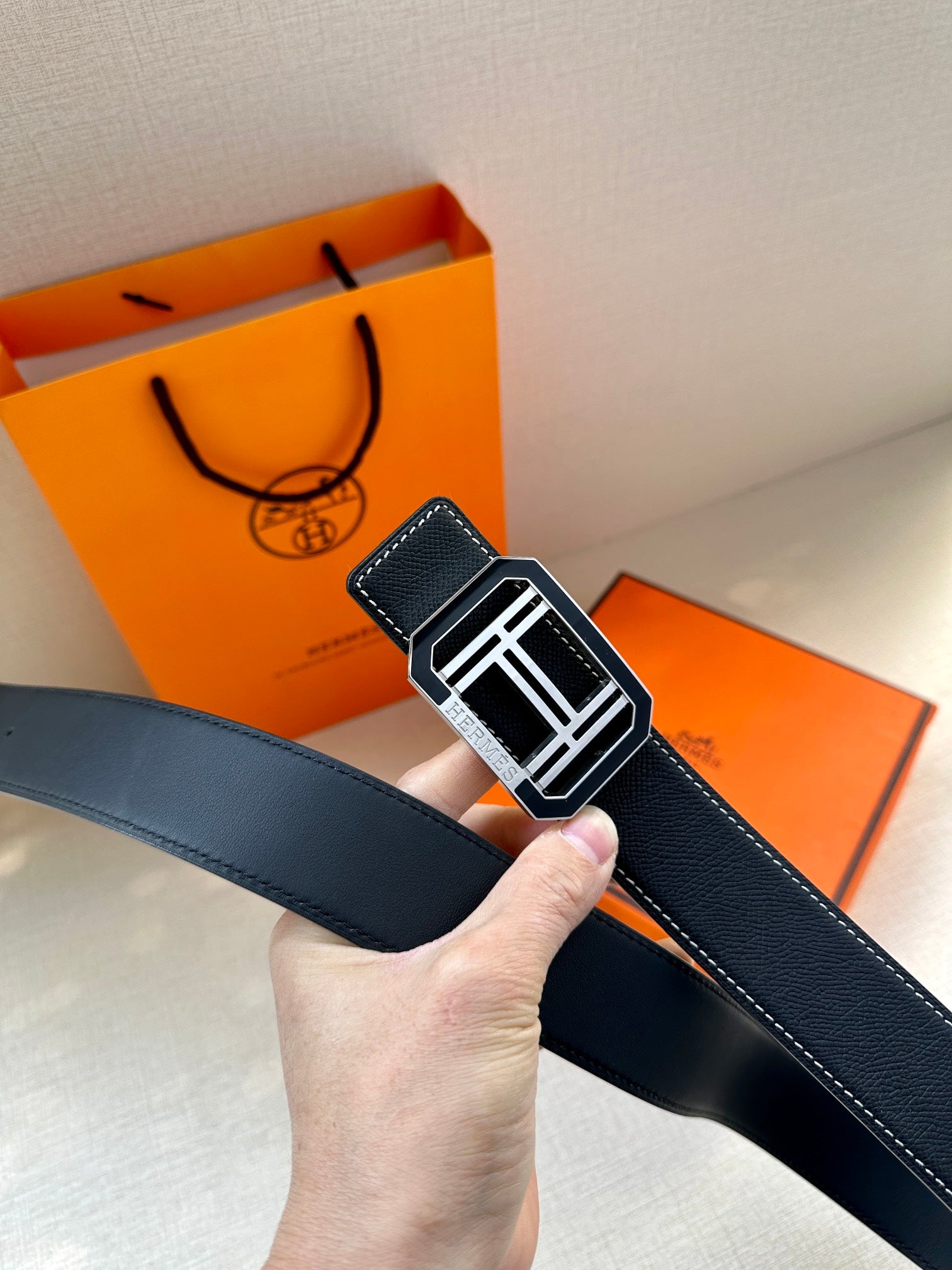 Dark leather belt held up, showing silver-tone square buckle and contrast white stitching.