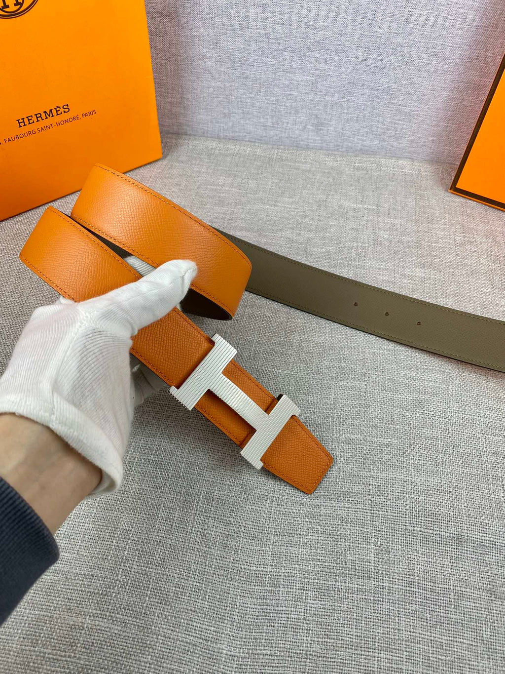 Two-tone leather belt with orange strap and white-gloved hand, H buckle close-up