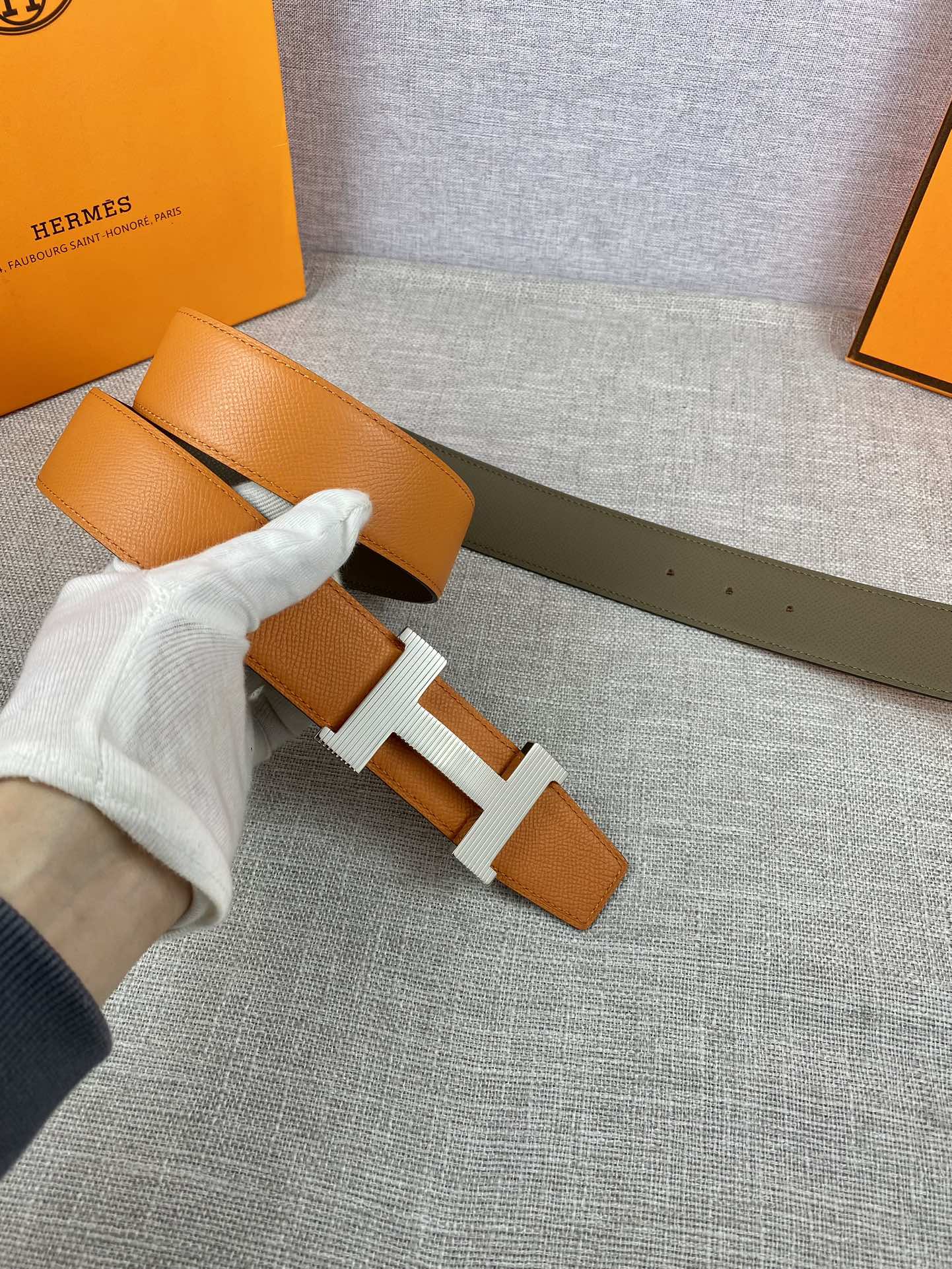 Two-tone leather belt with orange strap and white-gloved hand, H buckle close-up
