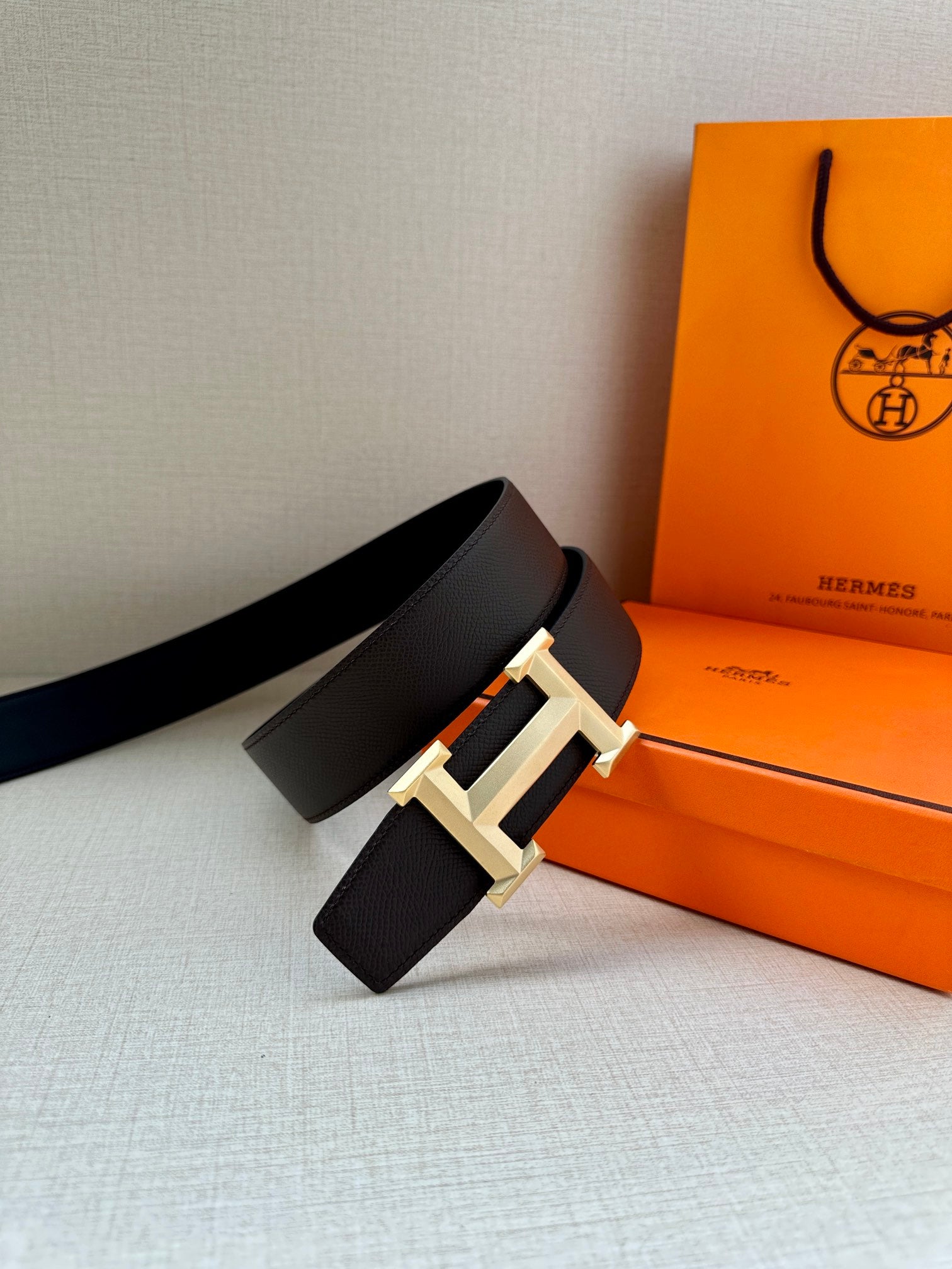 Leather belt with dark pebbled strap and gold-tone H buckle, laid on surface