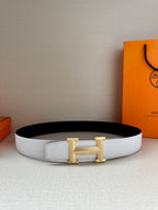 Wide leather belt with white strap and gold H buckle on a surface