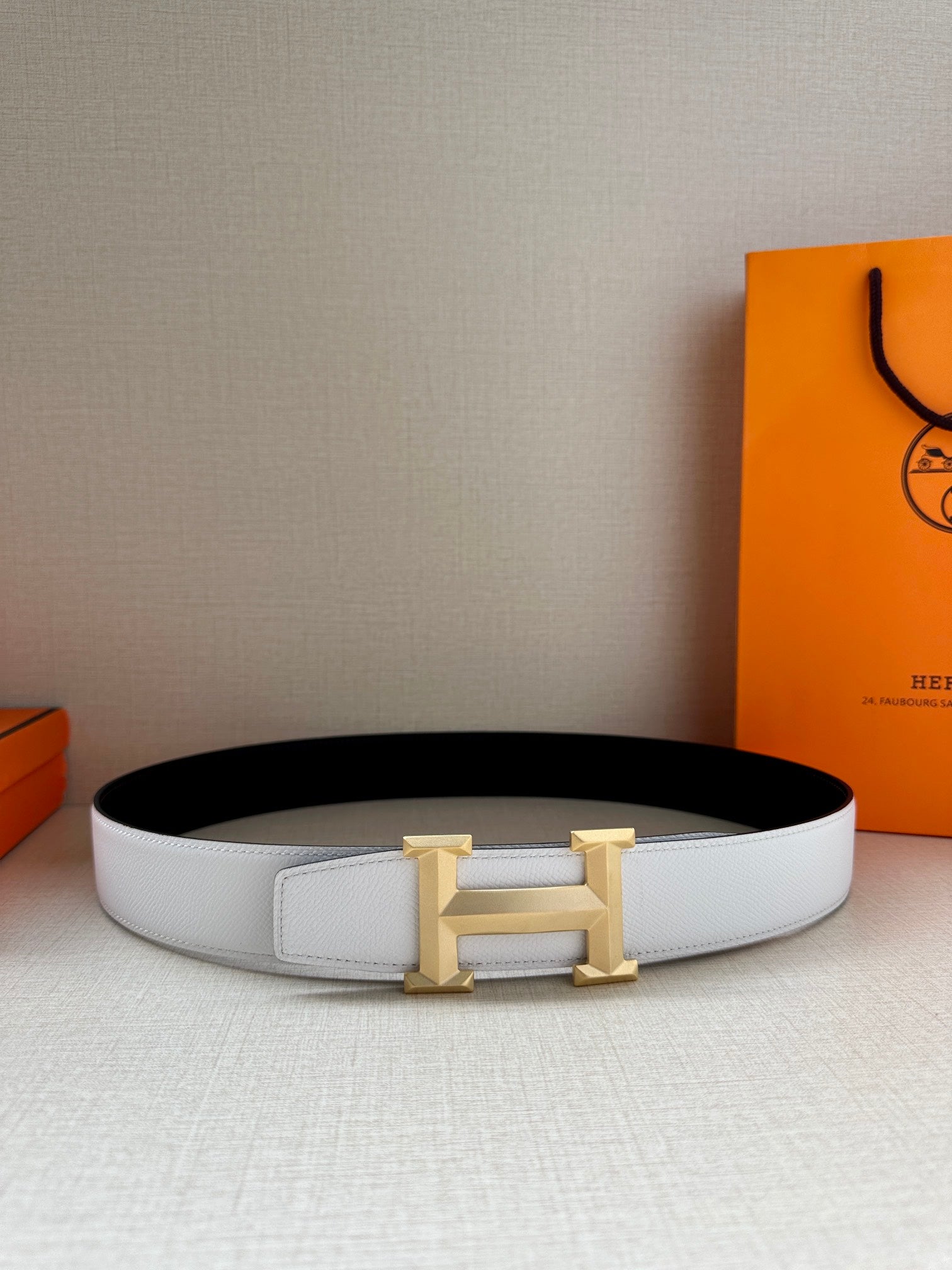 Wide leather belt with white strap and gold H buckle on a surface