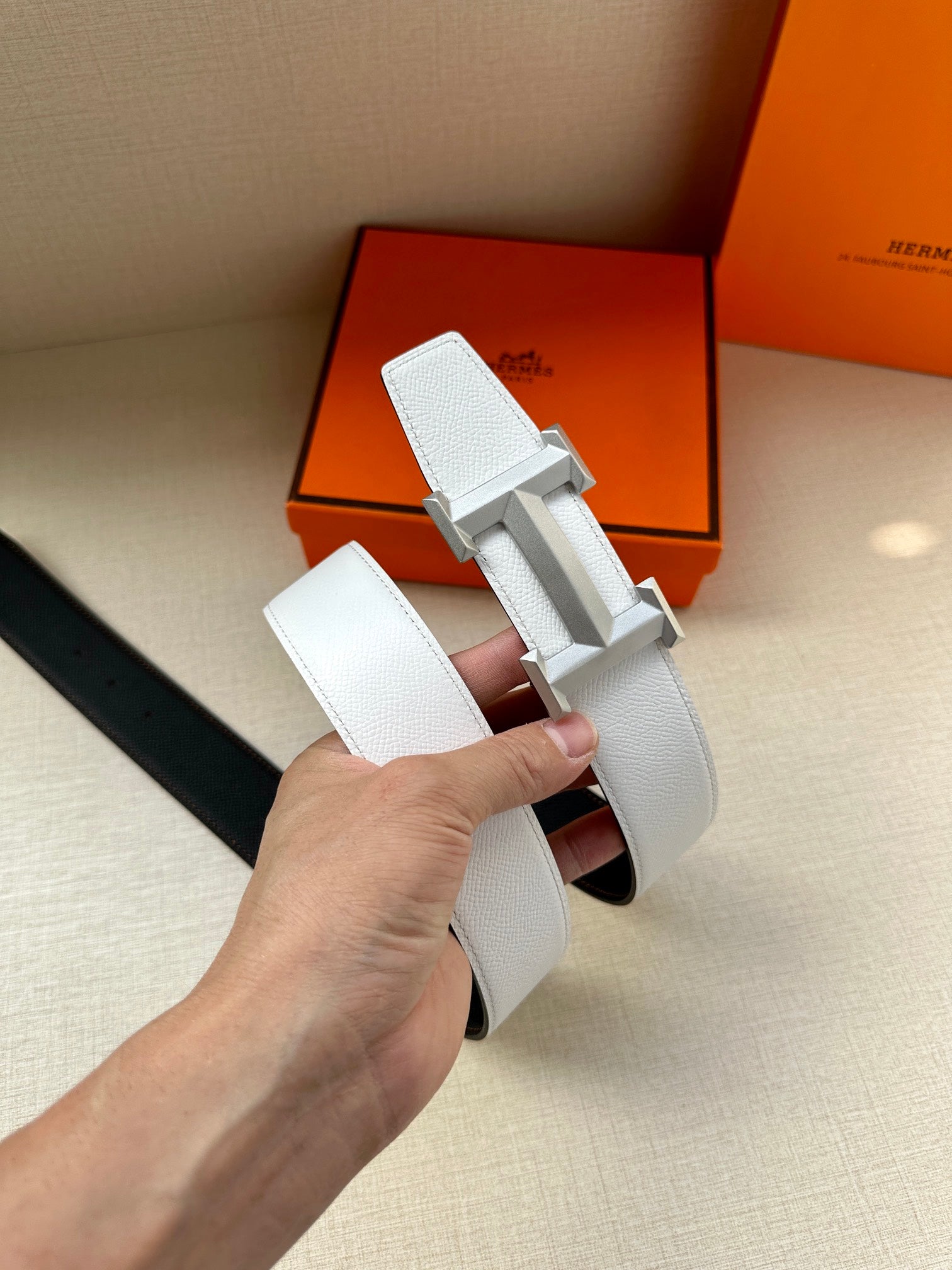 Leather belt with white strap and silver-tone H buckle, held in hand