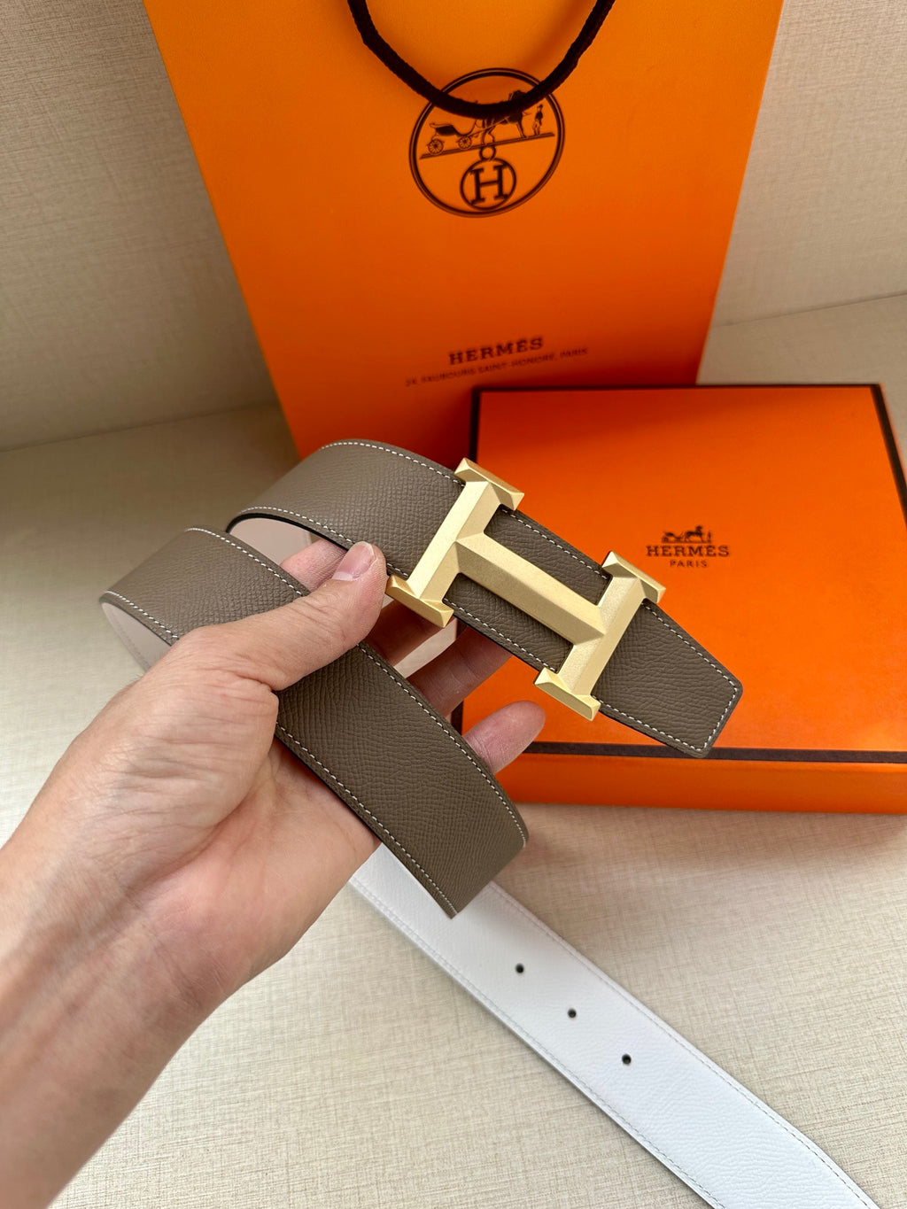 Hermes belt close-up held by hand: dark taupe strap with light gold-tone buckle
