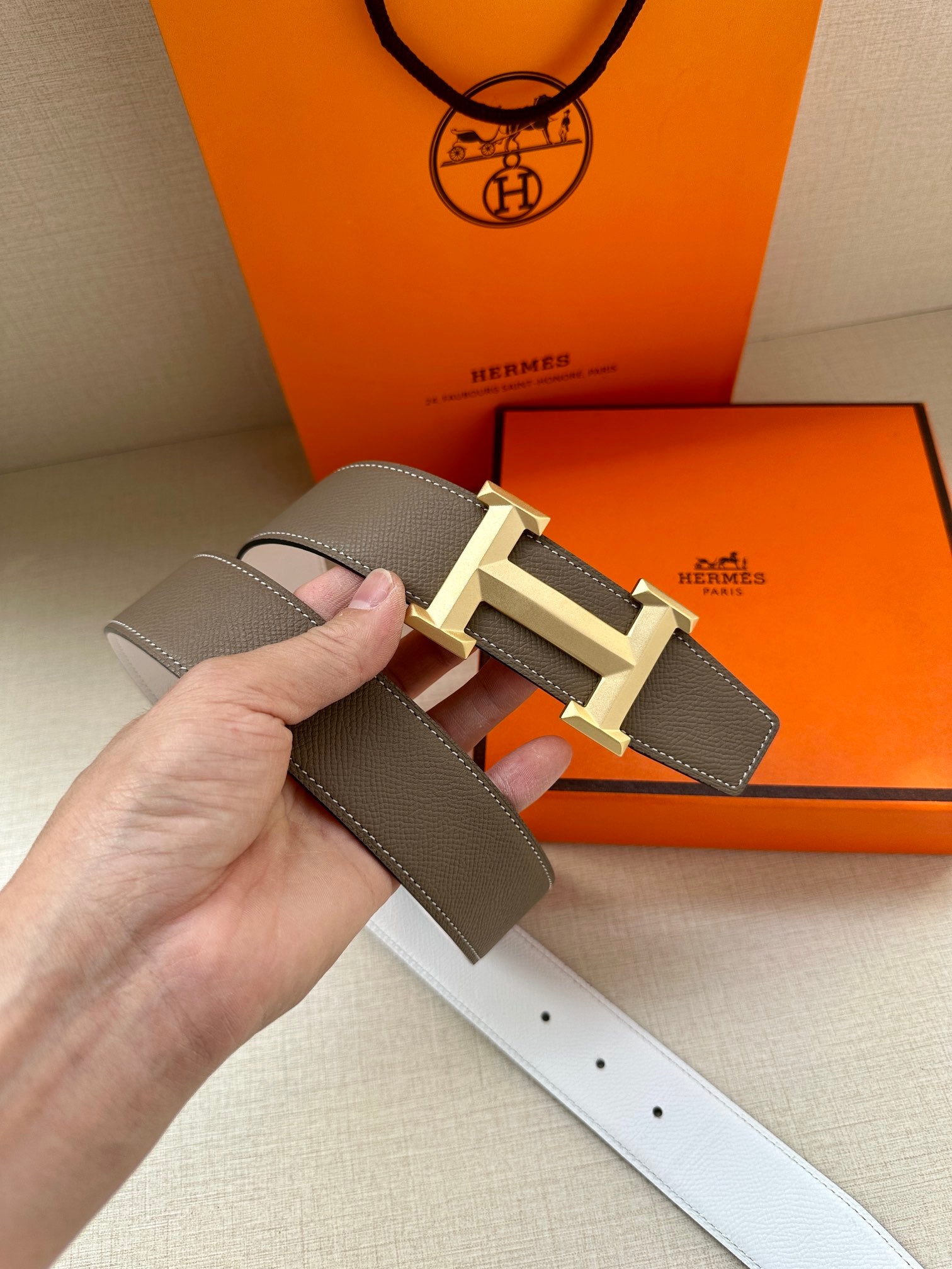Hermes belt close-up held by hand: dark taupe strap with light gold-tone buckle