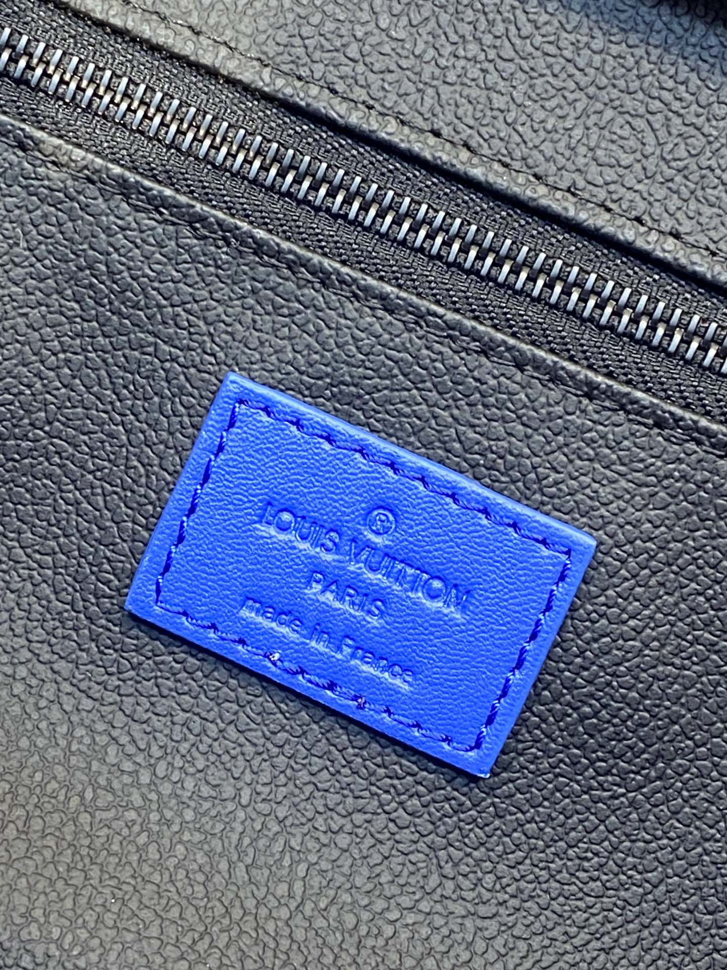 Blue Louis Vuitton embossed label on textured bag material near the zipper