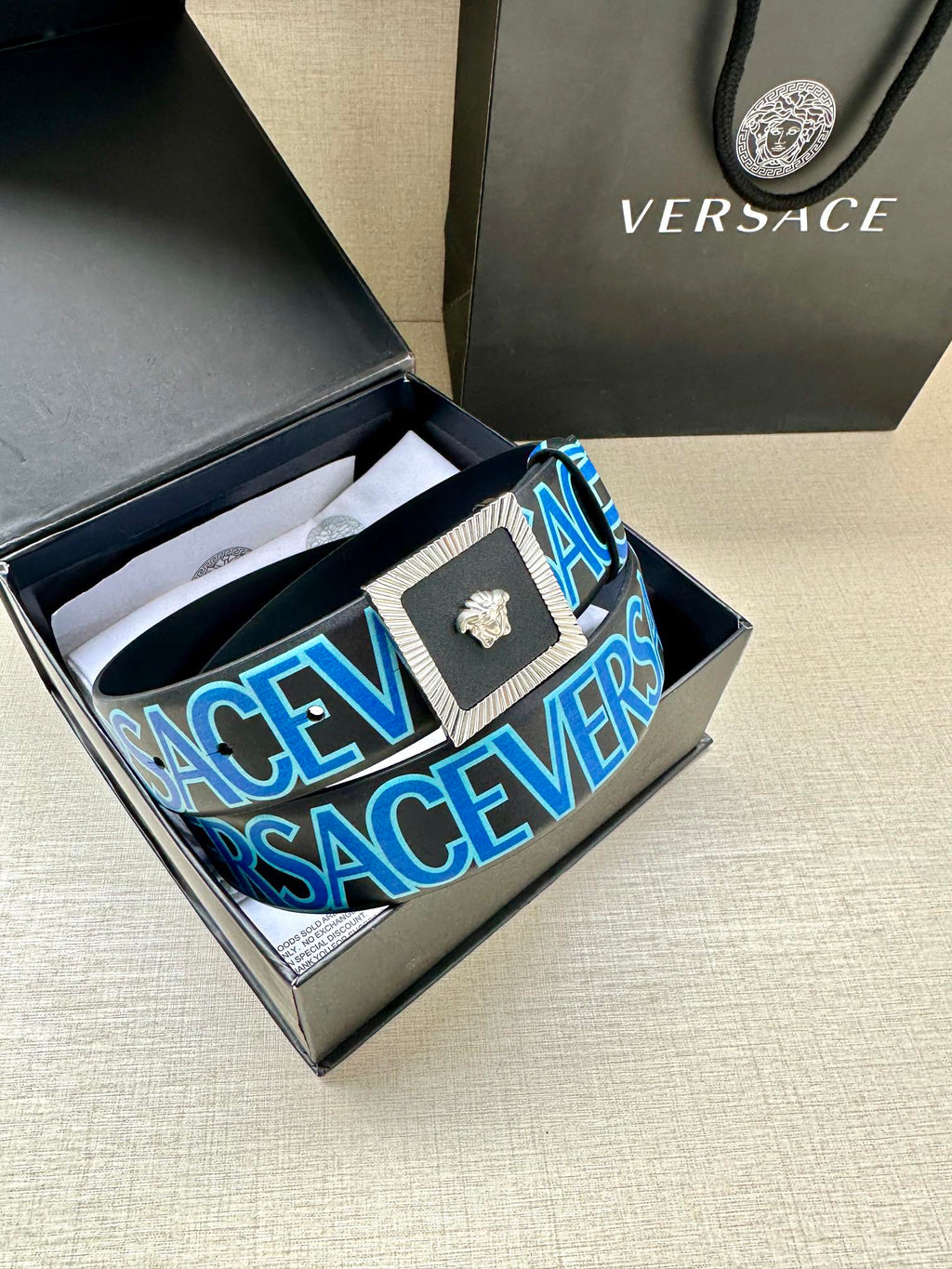 Versace logo belt inside box, blue strap with repeating lettering and gold buckle.