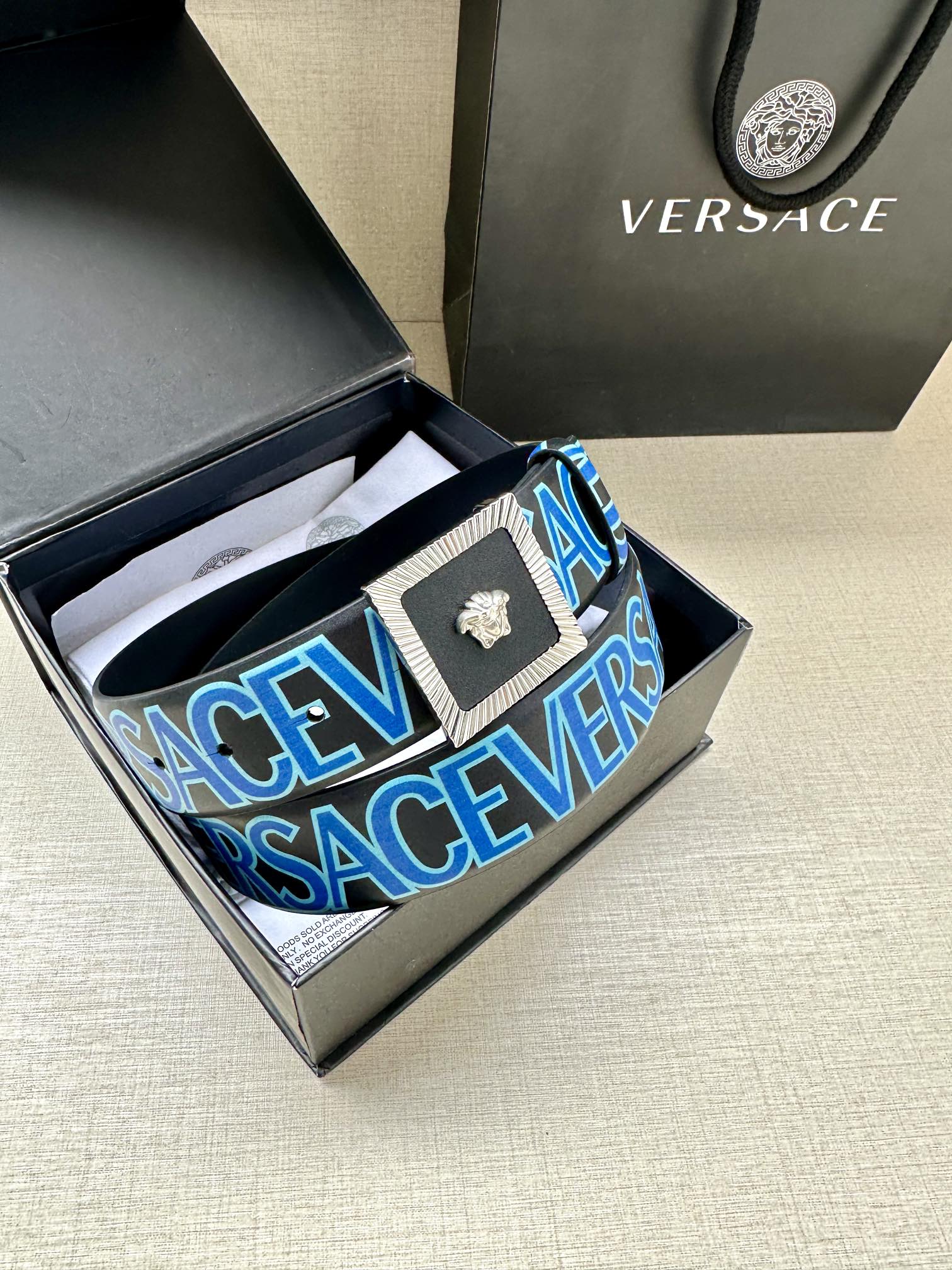 Versace logo belt inside box, blue strap with repeating lettering and gold buckle.