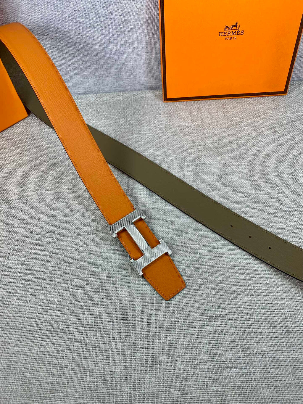 Orange leather belt with silver H buckle and olive keeper strap, laid flat