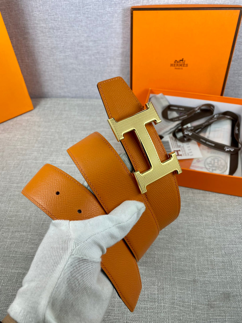 Orange leather belt with gold H buckle and olive strap, held in gloved hand