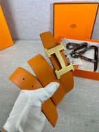 Orange leather belt with gold H buckle and olive strap, held in gloved hand
