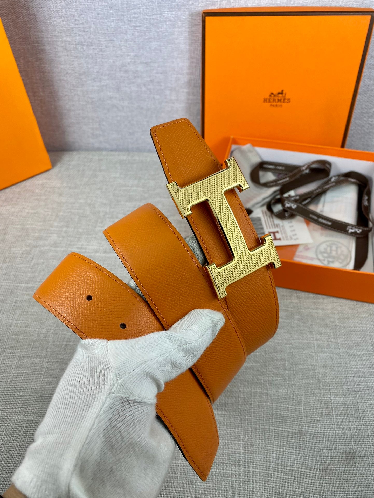 Orange leather belt with gold H buckle and olive strap, held in gloved hand