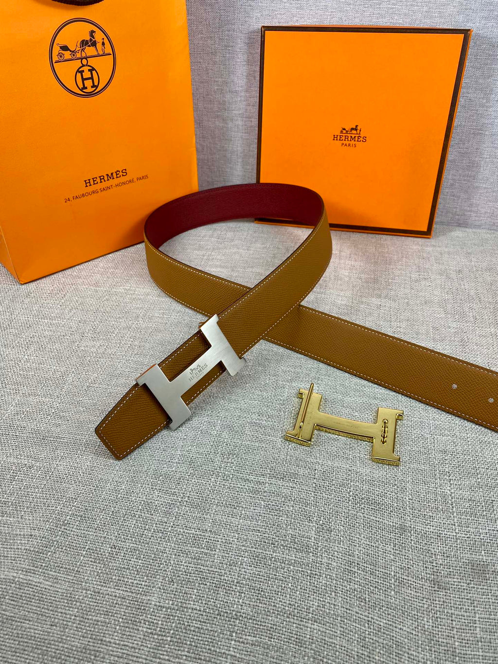 Leather belt with tan exterior, burgundy inner band, and gold H buckle detail.