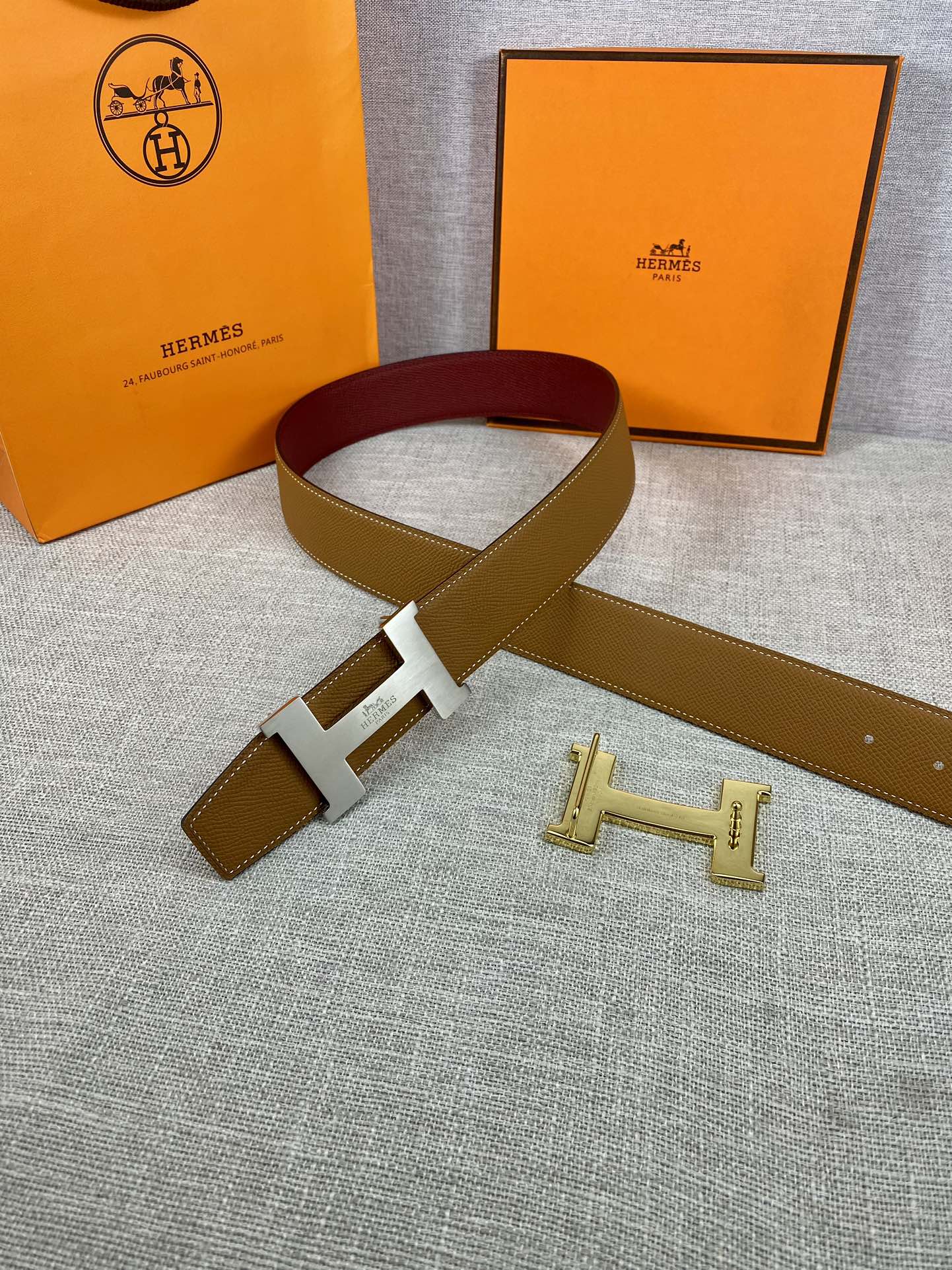 Leather belt with tan exterior, burgundy inner band, and gold H buckle detail.