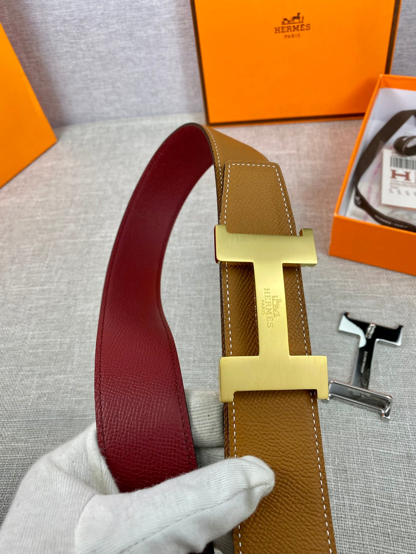 Leather belt close-up showing tan strap, gold H buckle, and burgundy inner band.