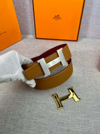Leather belt on hand, tan strap with gold H buckle and burgundy accent underside.