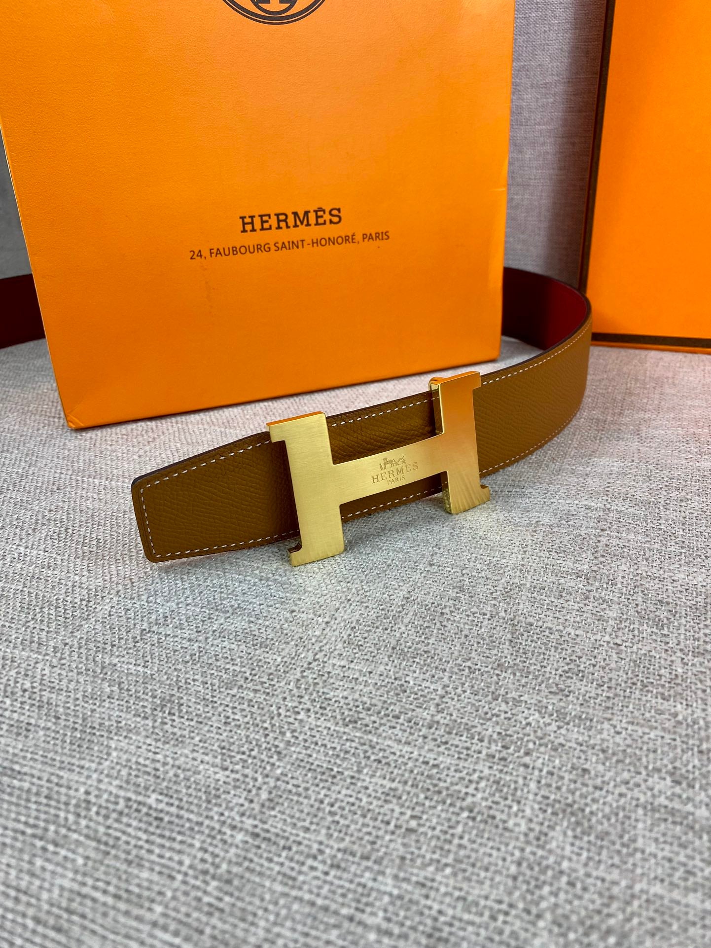 Leather belt with tan strap and gold H buckle, flat on a fabric surface.