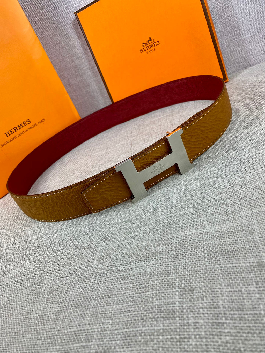 Leather belt with tan strap, gold H buckle, and burgundy inner band on display.
