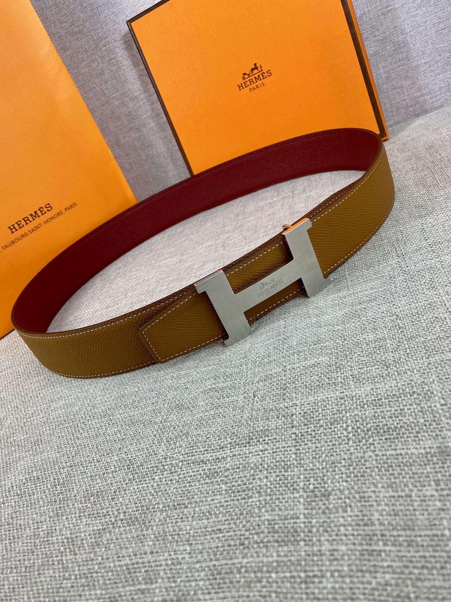 Leather belt with tan strap, gold H buckle, and burgundy inner band on display.