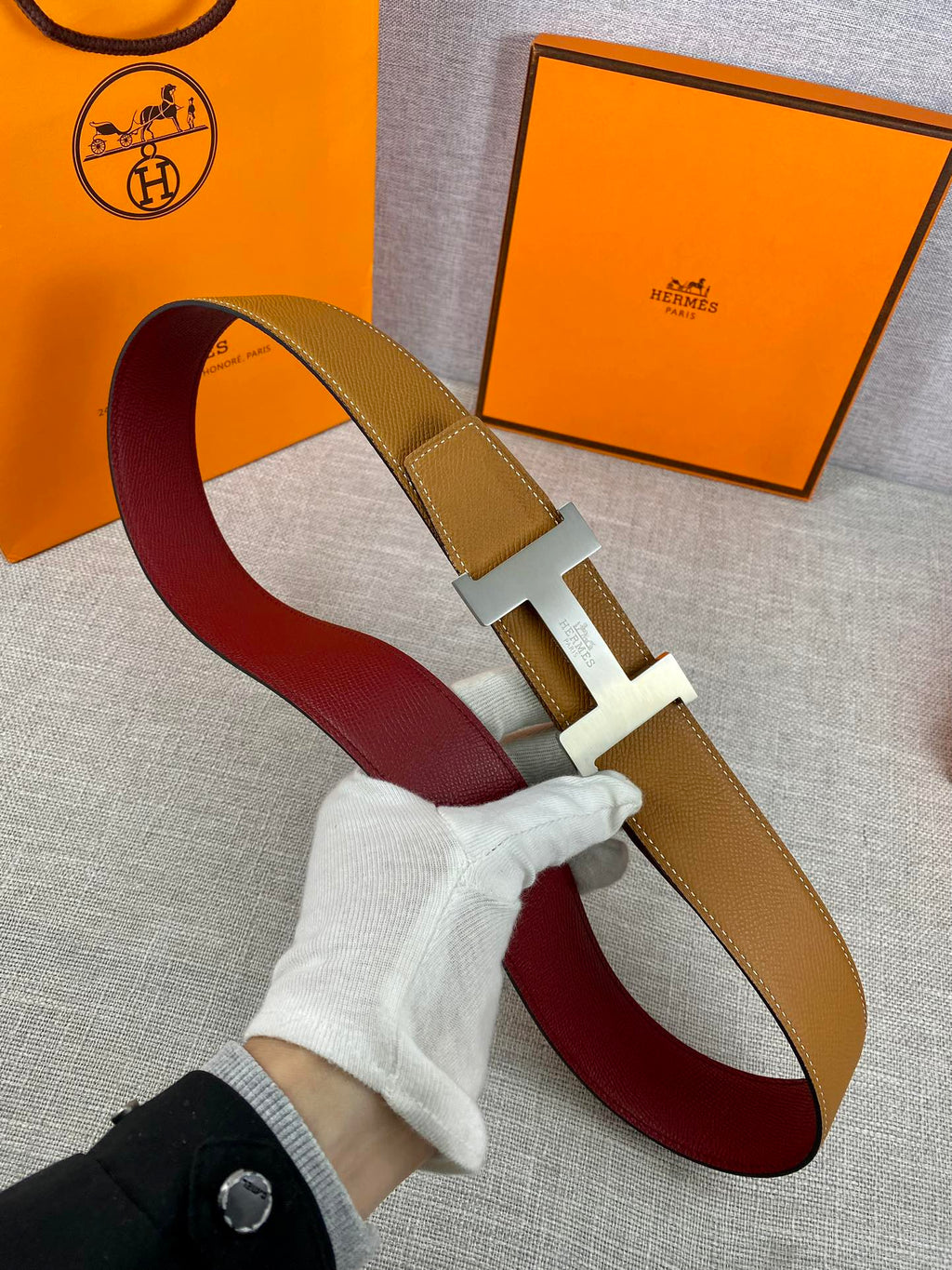 Leather belt with tan strap, gold H buckle, and burgundy underside looped on surface.