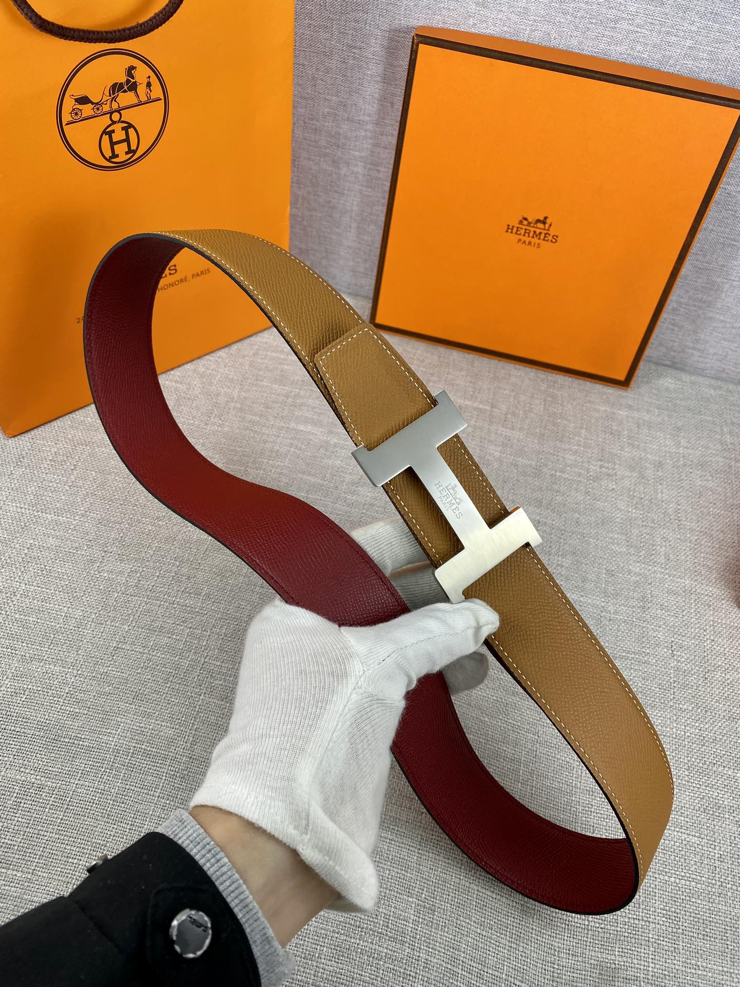 Leather belt with tan strap, gold H buckle, and burgundy underside looped on surface.