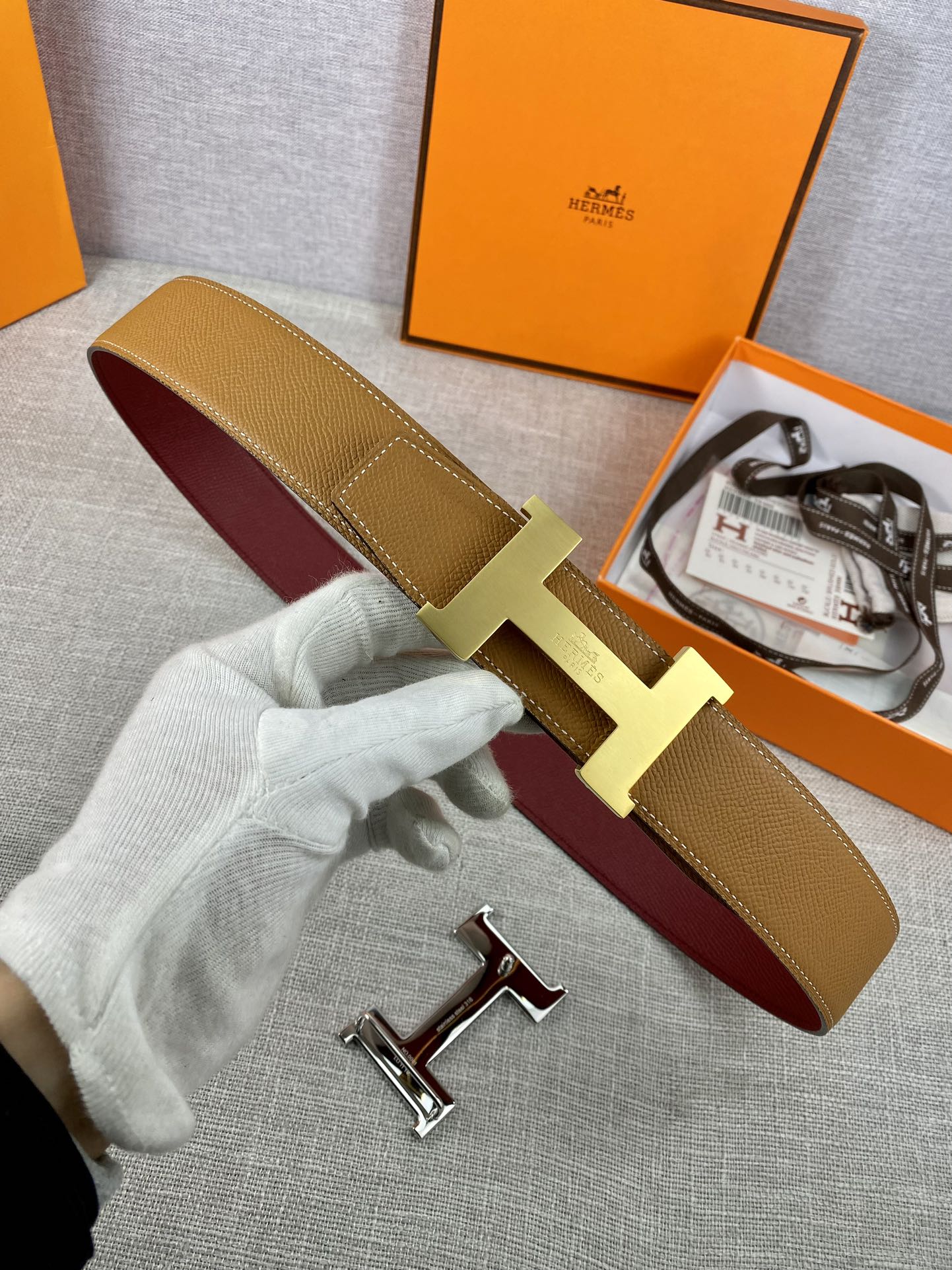 Leather belt with tan strap, gold H buckle, and burgundy underside with keeper.