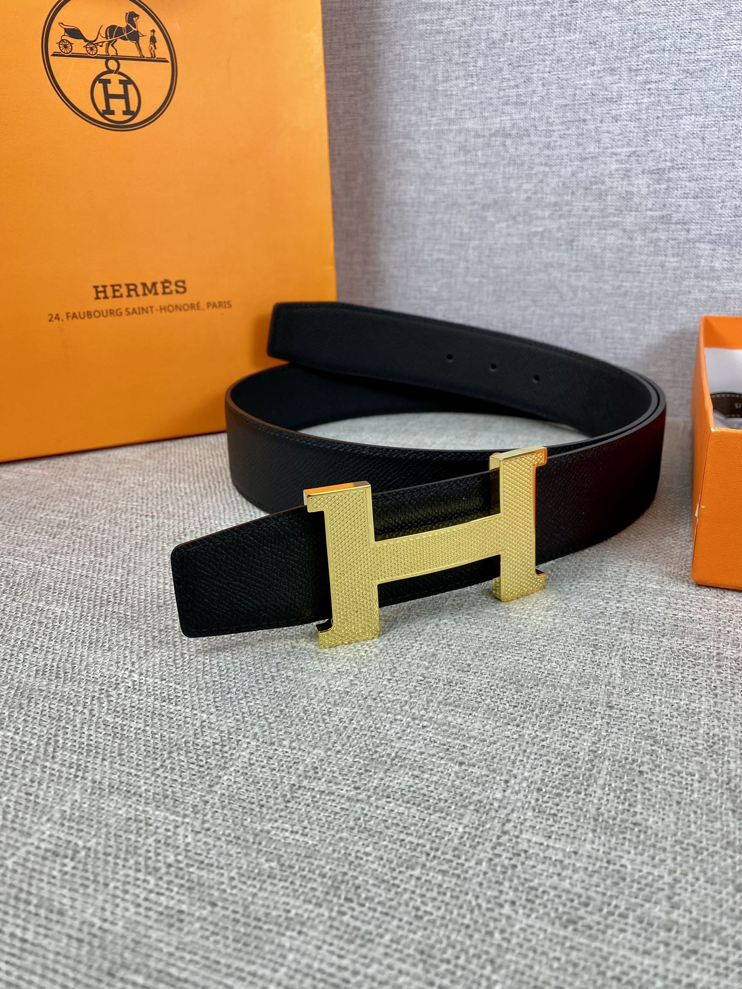 Black leather belt with gold-toned H buckle on a gray fabric surface