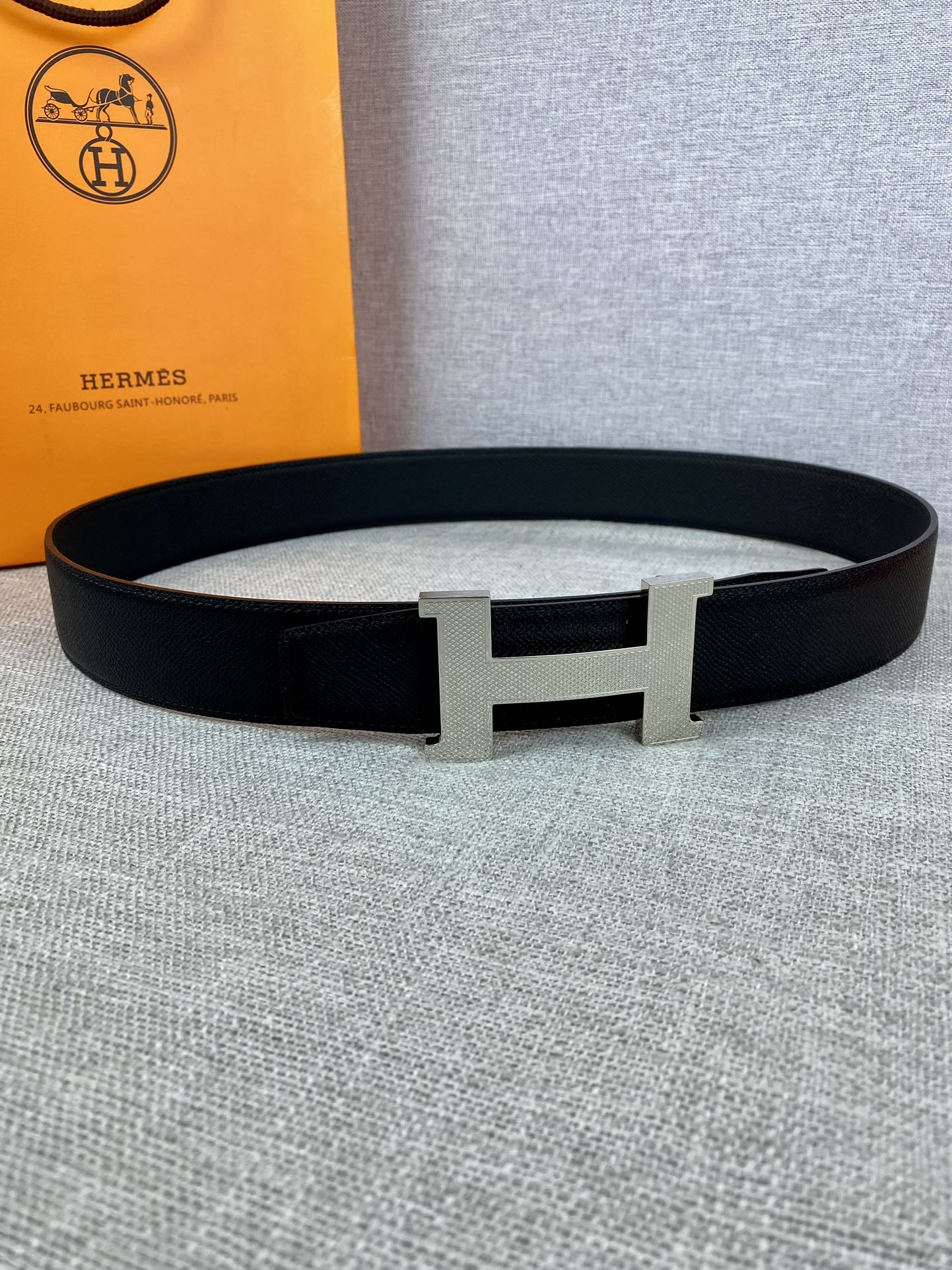 Black leather belt with silver-toned H buckle, displayed on gray fabric
