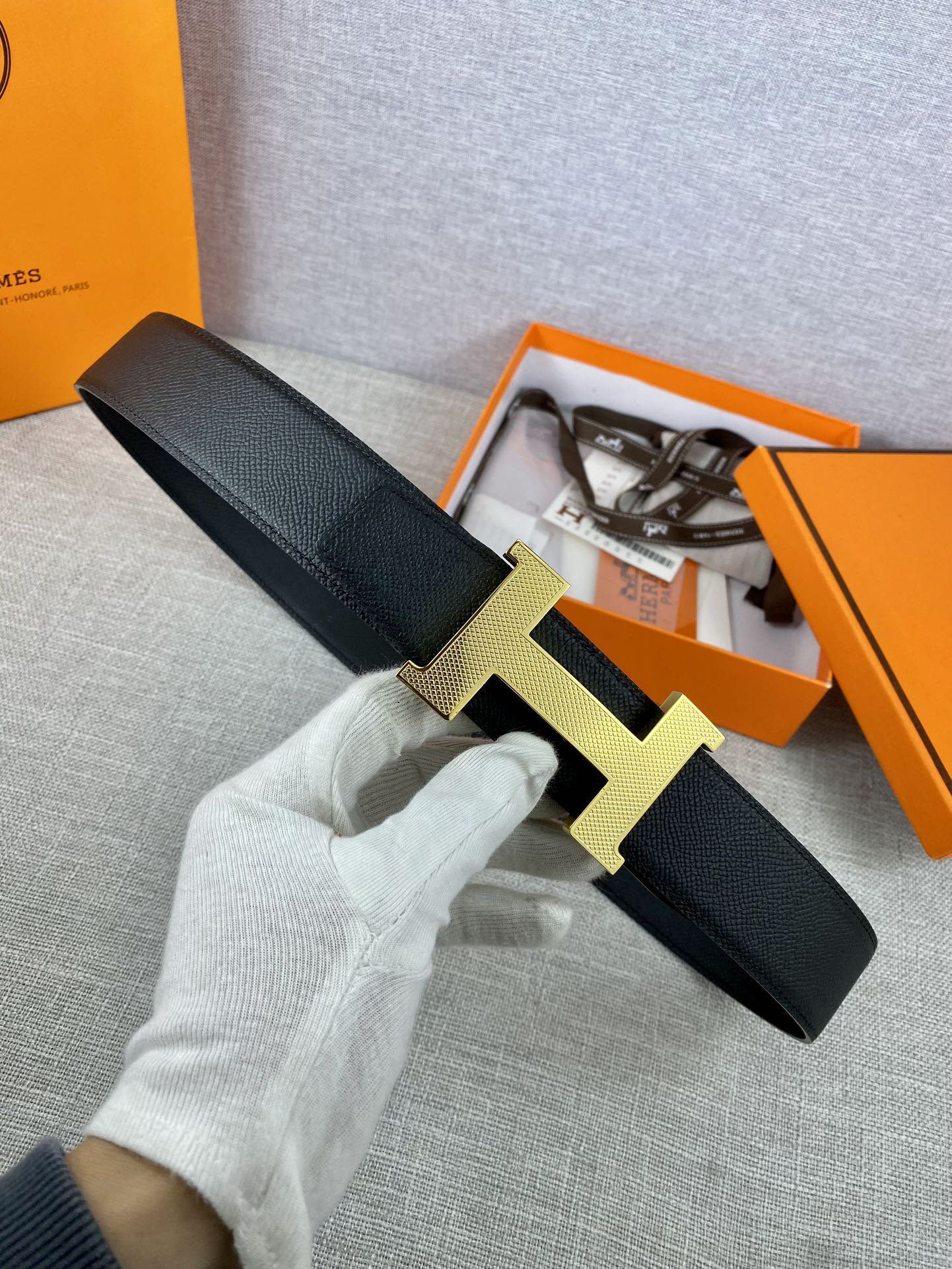 Black leather belt with textured gold-toned H buckle, laid on a gray fabric surface