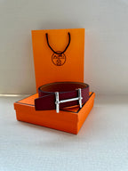 Leather belt with dark red strap and gold-tone double-prong buckle on orange box