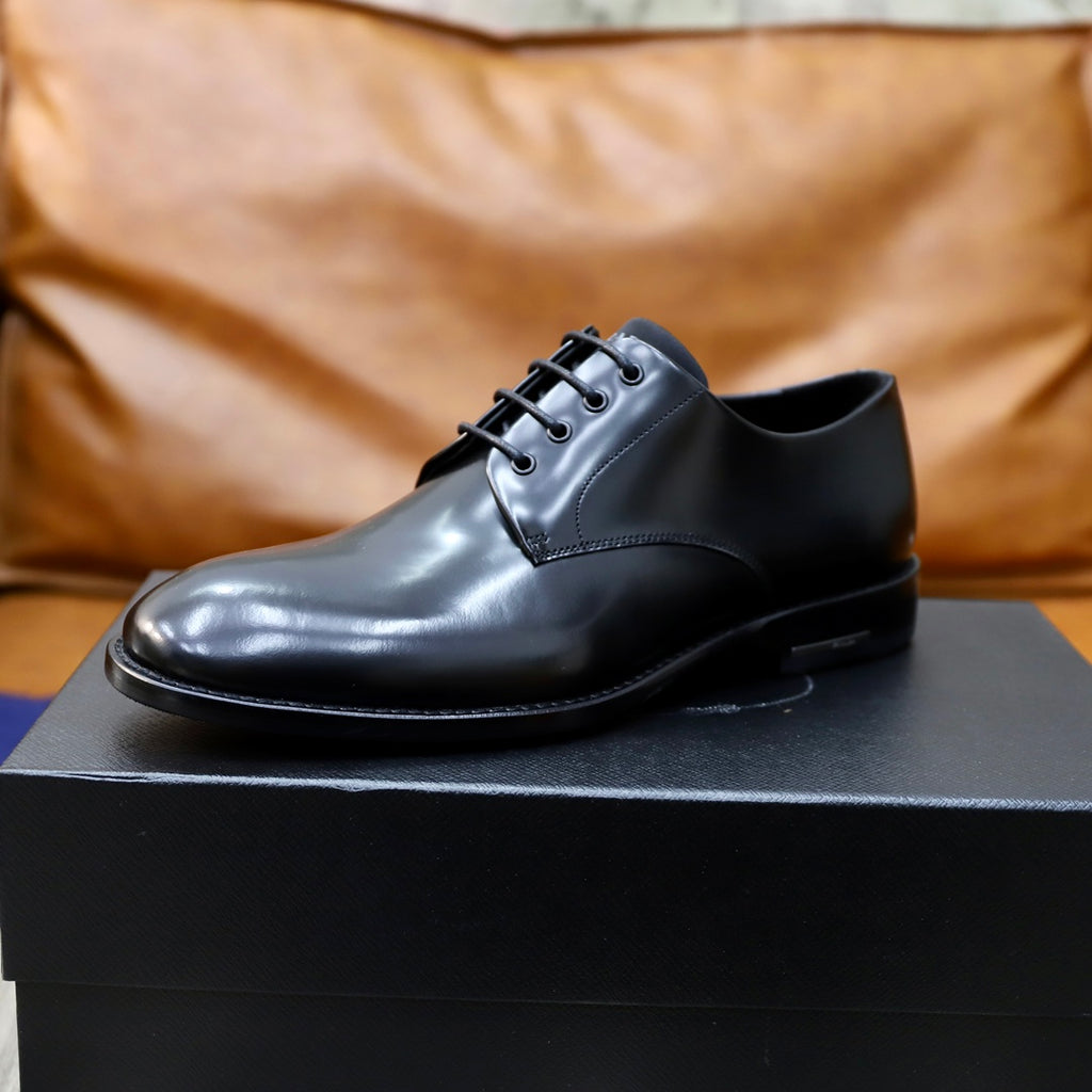 Prada unisex lace-up oxfords in dark shiny leather on black shoe box, front view.