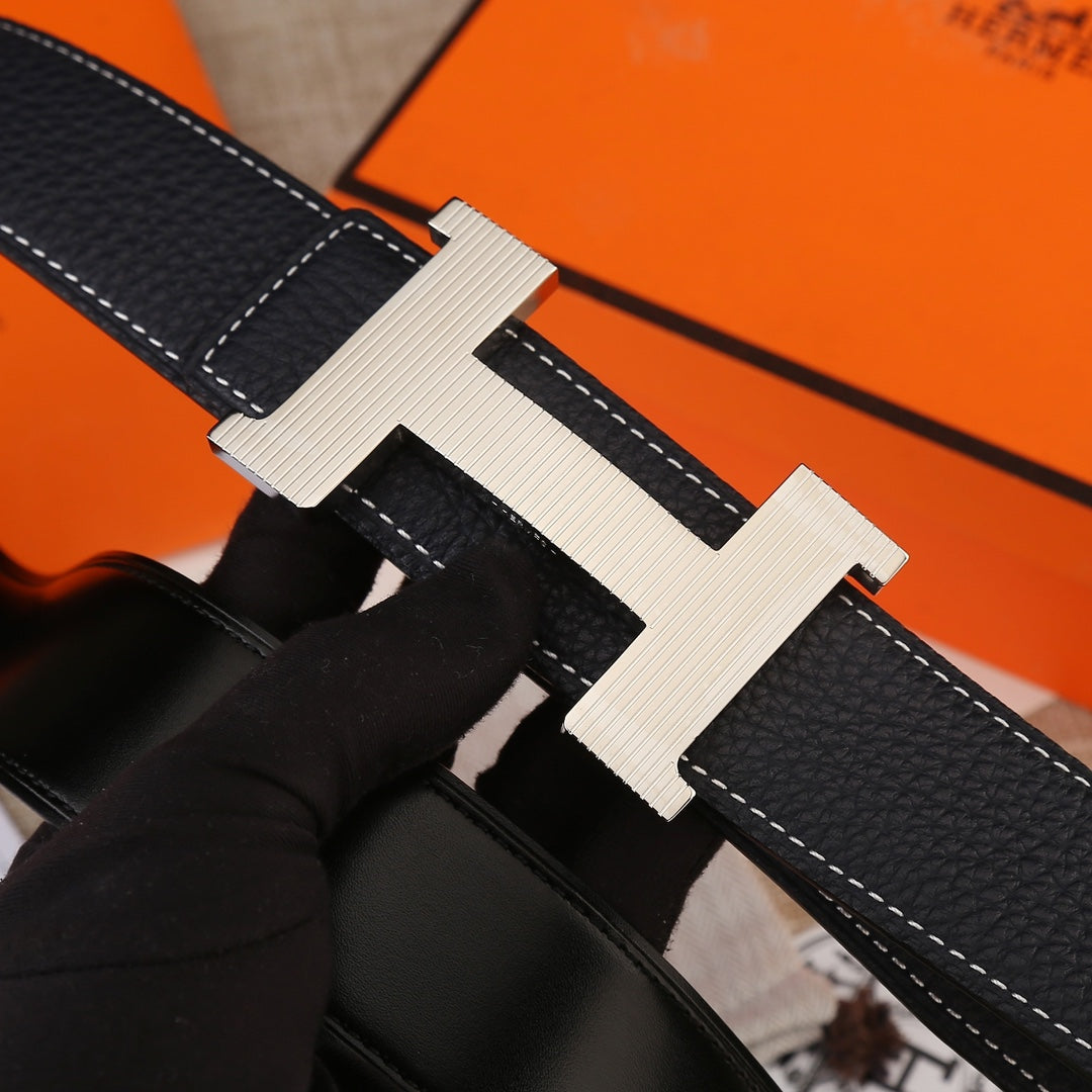 Hermes belt close-up of light gold H buckle on textured black leather with white stitching.