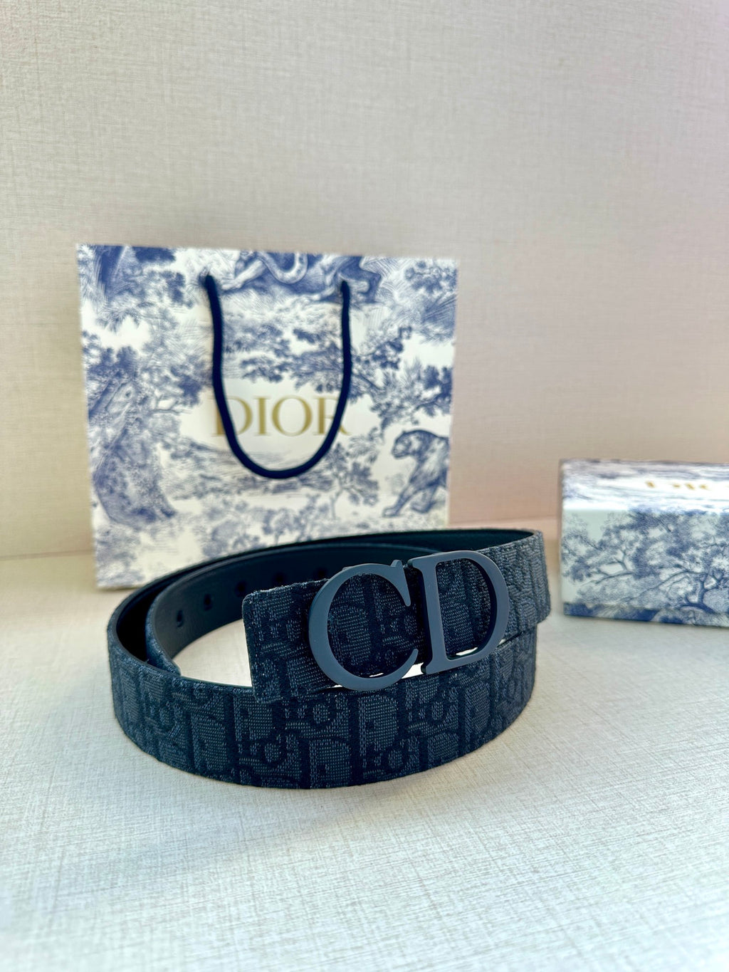 Dark blue designer belt with metal “D” buckle, placed on a box