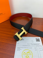Black leather belt with gold-tone H buckle and red inner lining, laid on table.