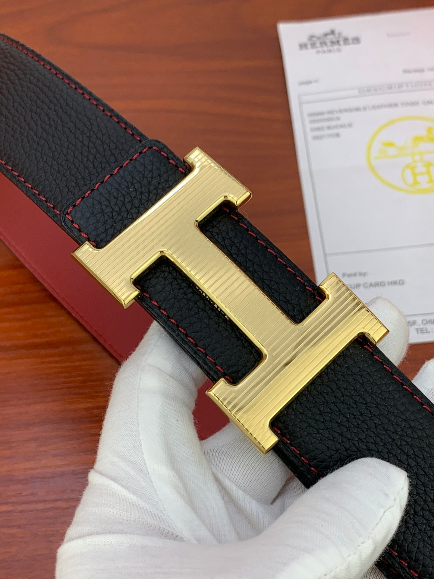 Black leather belt close-up with gold-tone H buckle and red edge stitching.